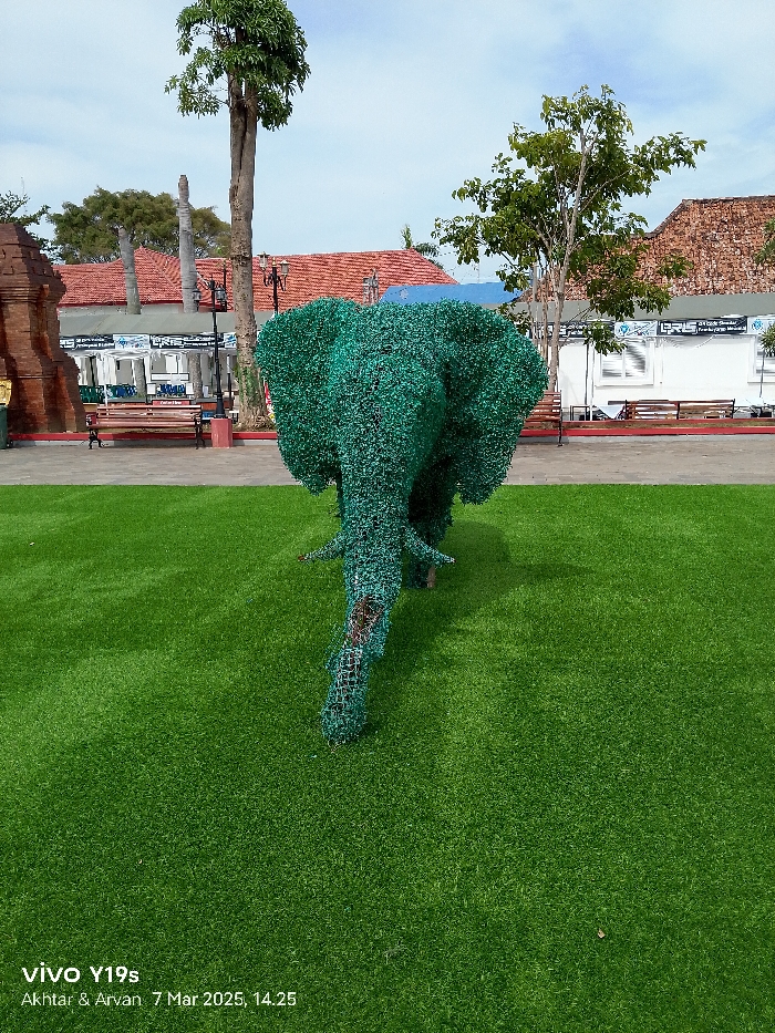 The image shows a green, wire mesh elephant sculpture standing on artificial grass. The sculpture is located in front of a red brick building with a white awning. The building is likely located in Indramayu, Indonesia, based on the provided coordinates (-6.327, 108.323). Two trees, one to the left and one to the right, are visible in the background. In the distance, there is a wooden bench and two lamp posts.