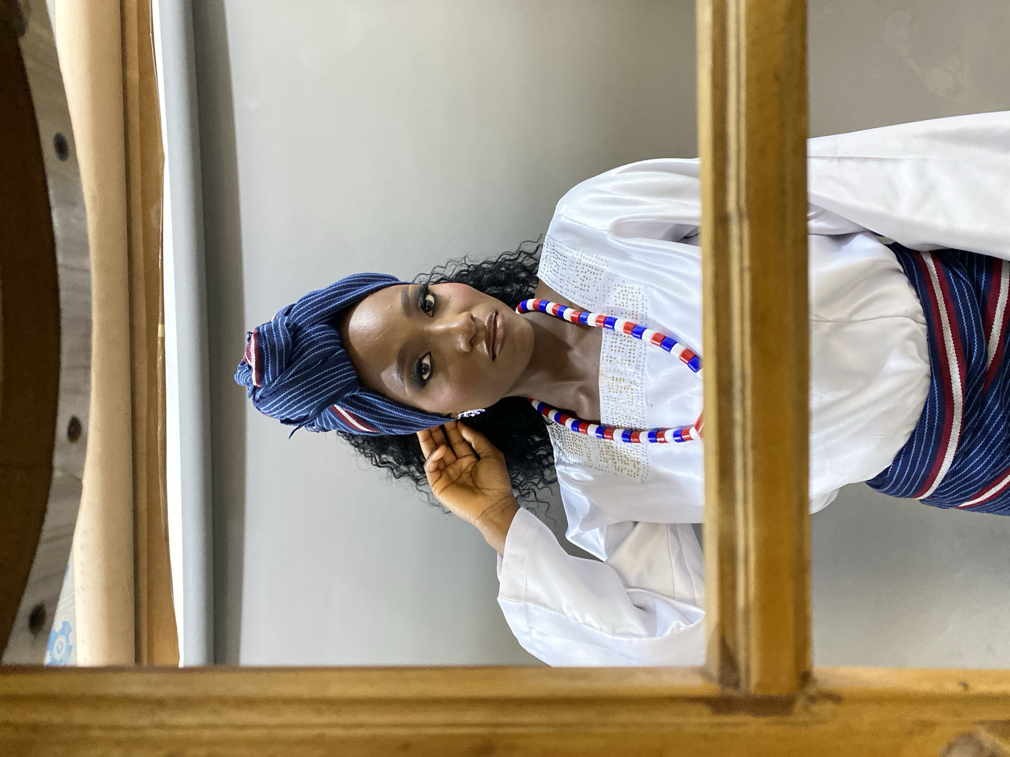 A medium shot shows a Black woman from the chest up, centered against a muted gray background. She is looking directly at the camera with a neutral expression. The woman wears a white long-sleeved garment with embroidered detailing around the neckline. A beaded necklace with alternating red, white, and blue beads rests on top of the garment. Her head is adorned with a blue and white striped headwrap, and her ears feature small, dangling earrings. Her dark, curly hair is visible peeking out from under the headwrap. A colorful striped sash, featuring dark blue, white, and red stripes, is visible around her waist. The image is framed by a wooden structure in the foreground, creating a window-like effect. The lighting is soft, illuminating the subject from the front. No specific details about the location in Gura Top, Nigeria, are discernible from the image itself.