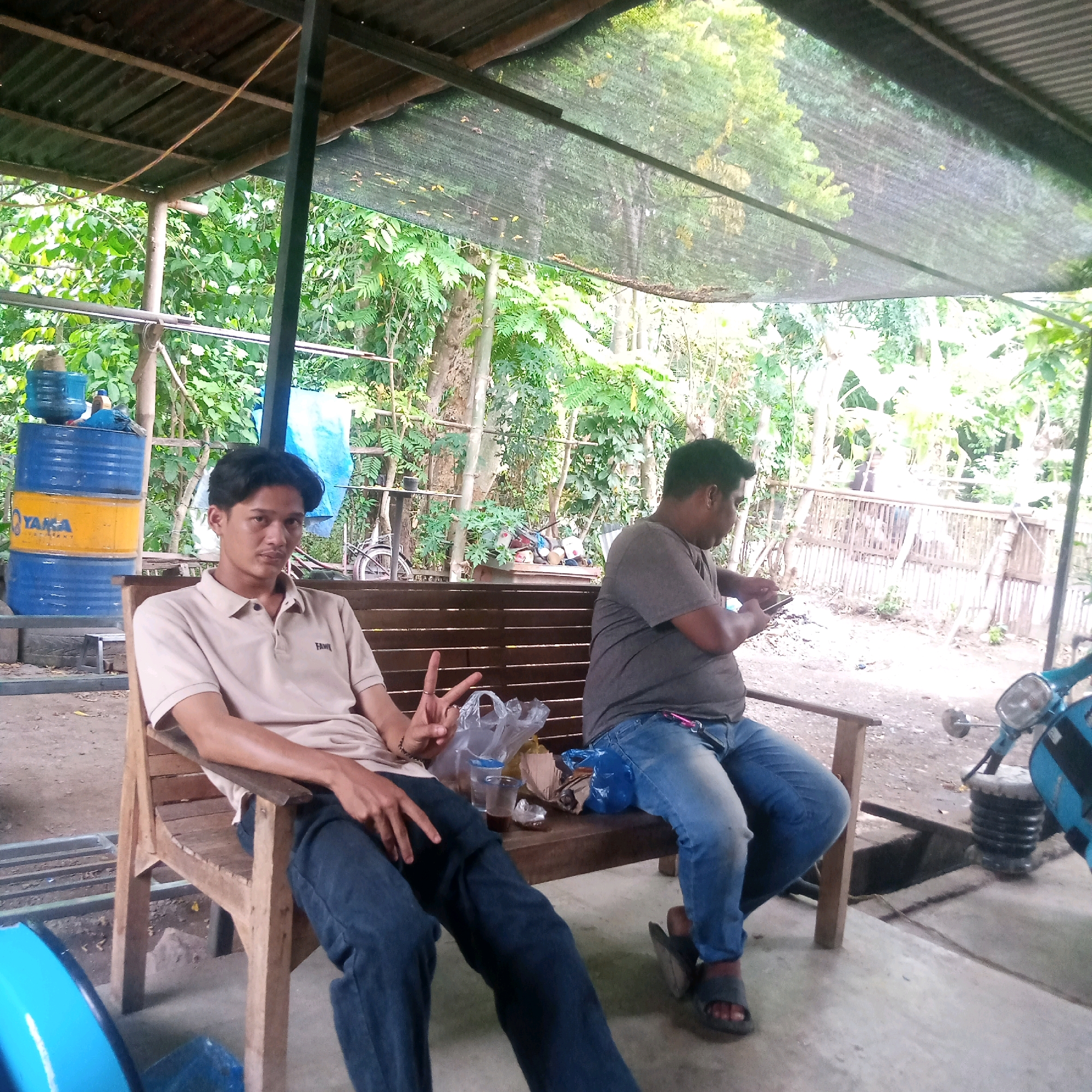 Two men are sitting on a wooden bench under a covered outdoor area. The man on the left is wearing a light brown polo shirt and dark blue jeans. He is looking at the camera and holding up two fingers in a peace sign. The man on the right is wearing a grey t-shirt and blue jeans. He is looking down at his phone and holding it in his hands. In front of them on the bench are two plastic cups, one with liquid in it, and a few plastic bags. To the left of the bench are two large blue and yellow barrels stacked on top of each other. A blue scooter is parked to the right of the bench. The background is filled with lush green trees and foliage. The ground is a mix of dirt and concrete.