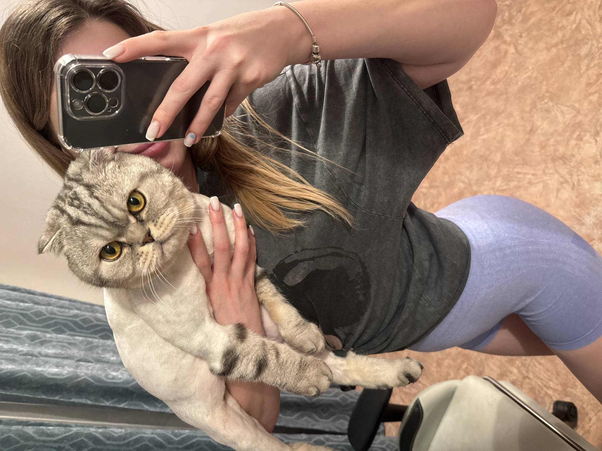 A person holds a gray and white cat while taking a selfie with a smartphone. The person is wearing a gray t-shirt, and light blue leggings. The cat has yellow eyes. The background is a textured wall.
