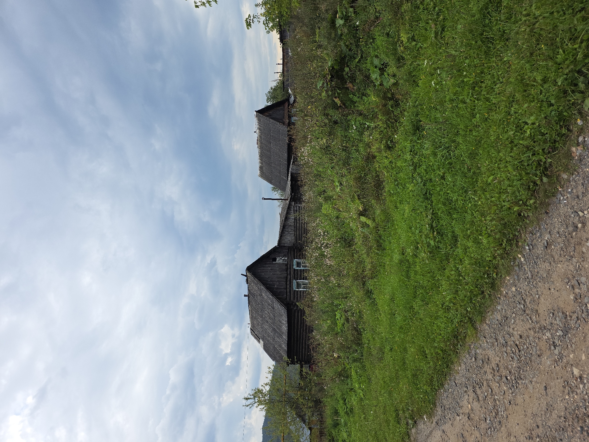 The image depicts a rural scene with two old wooden houses in the background, overgrown with vegetation. In the foreground, a dirt road is visible on the left, transitioning into a grassy area on the right. The sky is overcast with gray clouds. The houses appear to be constructed from logs and have weathered slate roofs. One house has two windows visible. The environment suggests a remote or undeveloped area, possibly a village. The location is identified as Magnitka, Russia.