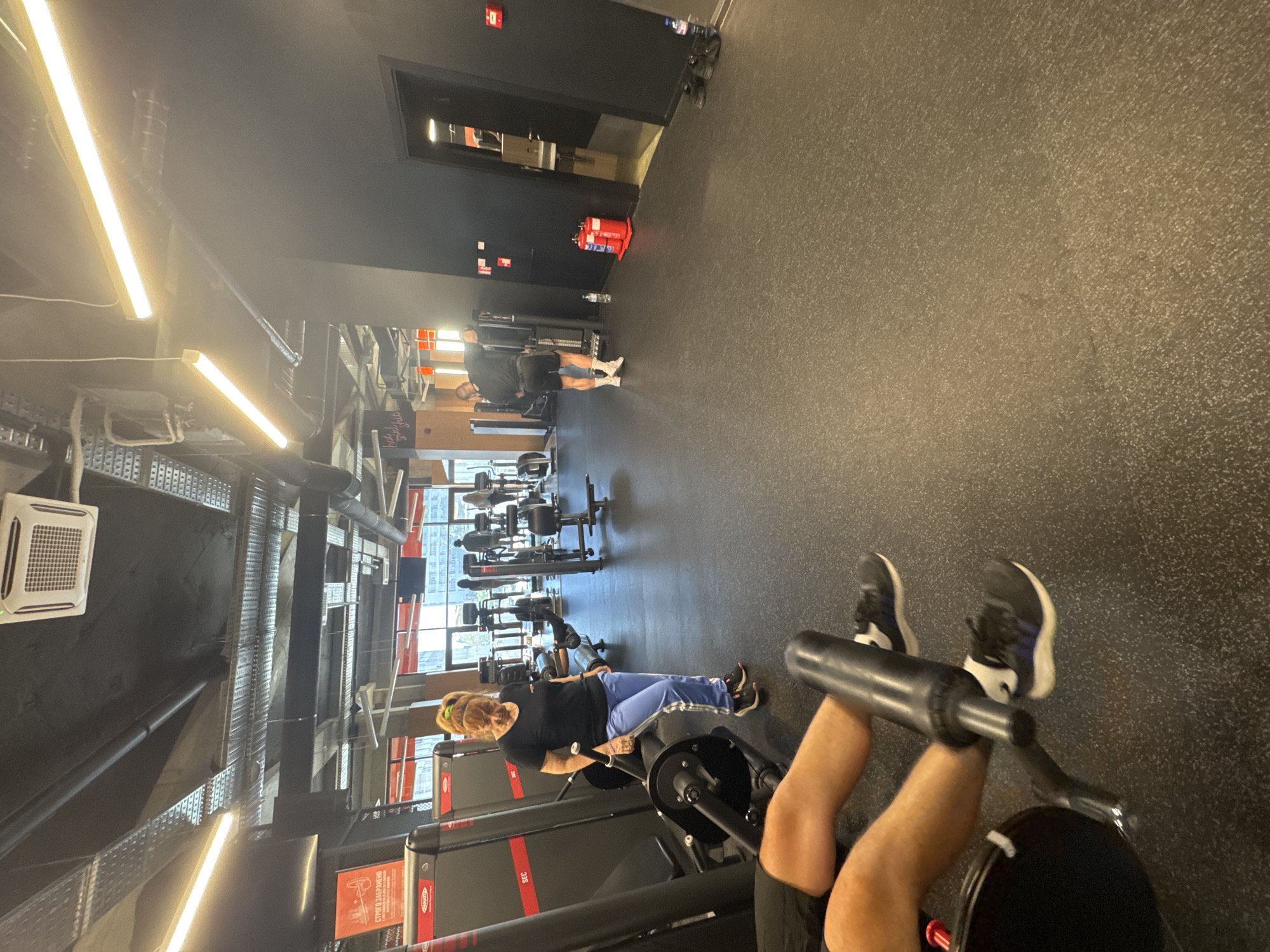The image shows the interior of a gym, likely in Plovdiv, Bulgaria, given the prompt's specification. The gym has black ceilings and a dark gray rubber floor. The foreground features a leg press machine with a person's legs visible using the equipment. Another person, a woman with blonde hair, is working out on a machine nearby. In the background, additional gym equipment is visible along with another individual exercising. The gym has windows offering an indistinct view of the city. The lighting is provided by overhead fluorescent lights. A fire extinguisher and other equipment are placed along the walls.