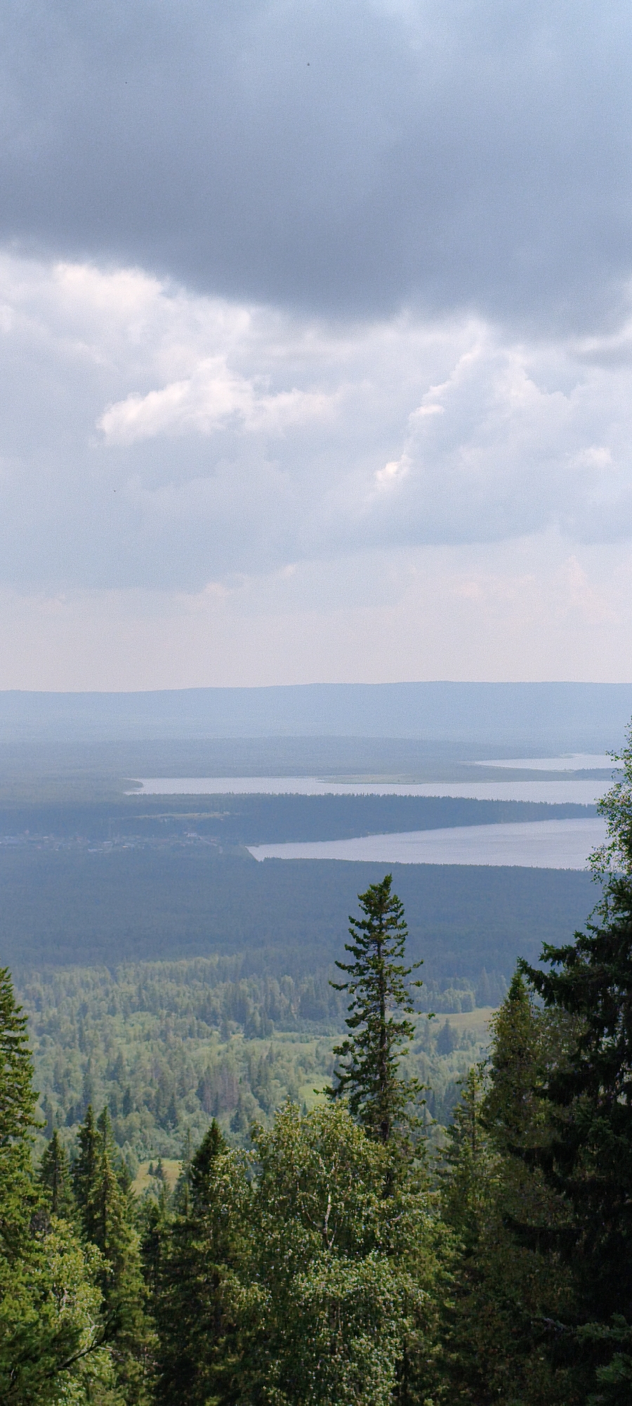 The image depicts a panoramic view from a forested elevation overlooking a vast expanse of natural landscape. In the foreground, dense evergreen and deciduous trees dominate the frame, with tall pine trees prominent. The middle ground reveals a large, dark green forest that stretches towards a body of water. Beyond the forest and water, layers of hazy, blue-hued mountains fade into the distance under a sky filled with substantial, textured clouds. The scene is likely taken from a natural viewpoint such as a mountain or hill in or near the city of Ziuratkul', Russia, known for its national park and lake. There are no people or specific objects clearly discernible, only the natural environment.