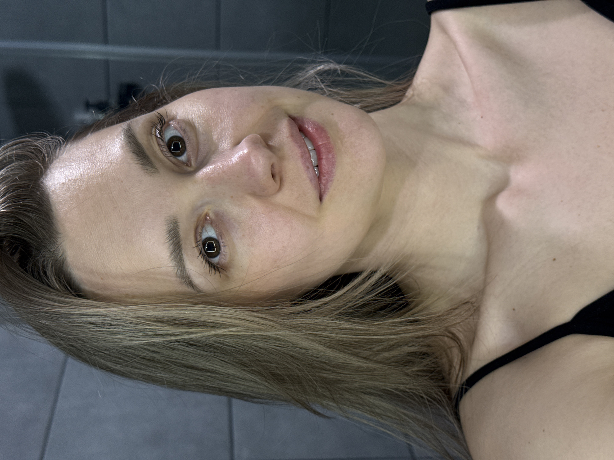 A close-up, rotated portrait photograph depicts a fair-skinned woman with brown eyes and light brown hair. She is wearing a black top with thin straps. The background appears to be a gray wall with horizontal tile lines, possibly a bathroom. There is no information to indicate the image was taken in Chaikovsky, Russia.