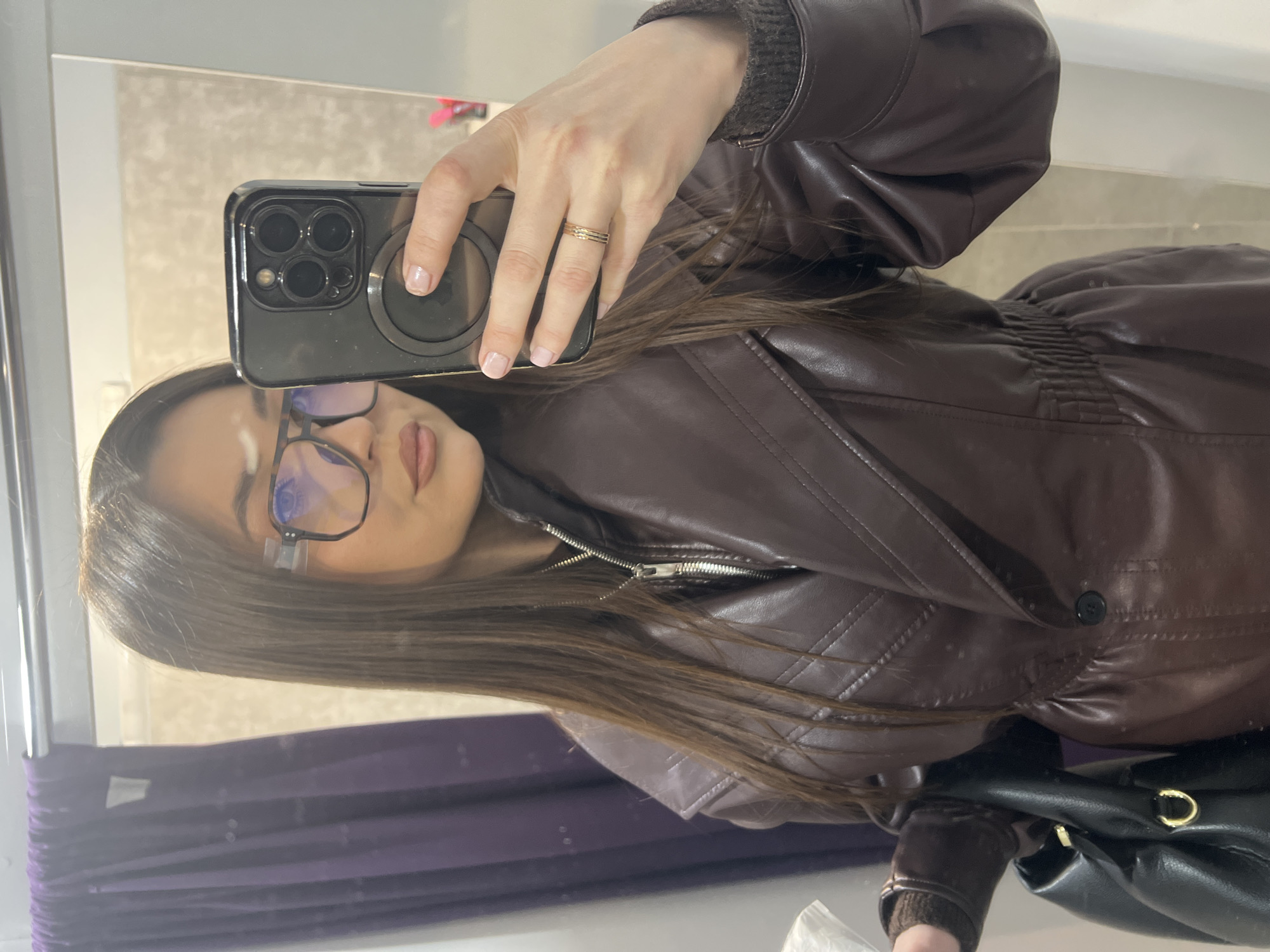 A woman with long brown hair and glasses is taking a selfie in a mirror. She is wearing a brown leather jacket with a zippered front. Her phone is a dark color with three camera lenses. She has a ring on her finger. In the background, there is a purple curtain. The image was likely taken indoors. There is no information available to determine if the location is in Khimki, Russia.