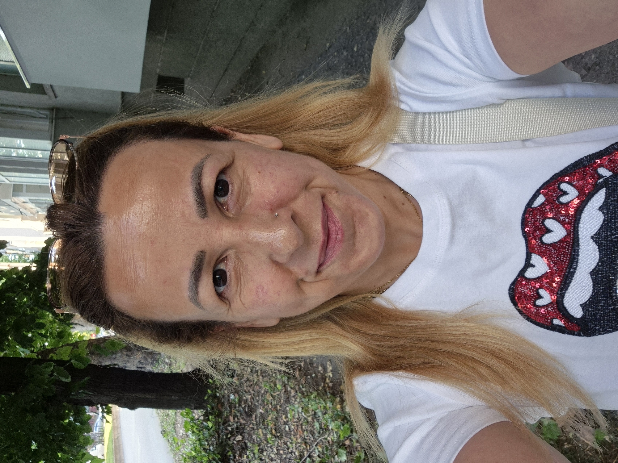 A woman is taking a selfie outdoors in front of a building with a tree to her left. She is looking at the camera and smiling. She has blonde hair and is wearing a white t-shirt with a sequined lip design. Her sunglasses are pushed up on her head. The background shows a sidewalk, a street, and a building.