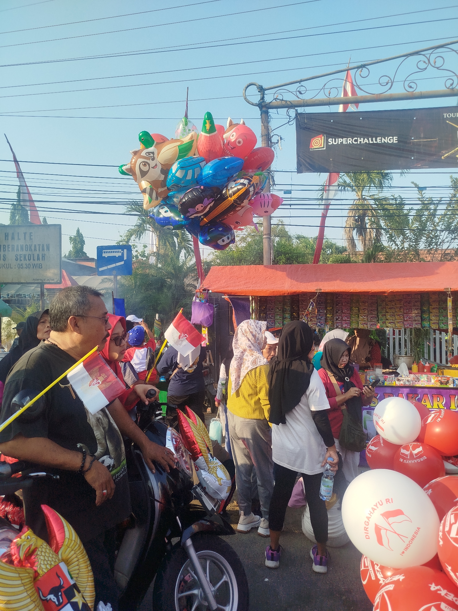 This image captures a street scene in Burneh, Indonesia, likely during a festive or commemorative event, as suggested by the abundance of red and white balloons, flags, and banners. In the foreground, a group of people are gathered around a motorcycle. A man in a black shirt and glasses holds a small Indonesian flag. Beside him, a woman in a red headscarf also holds an Indonesian flag. The motorcycle is decorated with more Indonesian flags and balloons featuring the Garuda Pancasila emblem, the national symbol of Indonesia.
Behind the motorcycle, a makeshift stall is set up with an orange canopy, selling various items that appear to be snacks or small goods. Several people, mostly women wearing headscarves, are interacting at the stall, some looking at their phones. In the background, more people are visible, and a banner with the text "SUPERCHALLENGE" is displayed above the stall. A sign with Indonesian text, possibly indicating a bus stop or school route, is also present. The overall atmosphere suggests a public gathering with patriotic undertones, possibly celebrating Indonesian Independence Day or a similar national holiday. The sky is clear and blue, indicating fair weather.