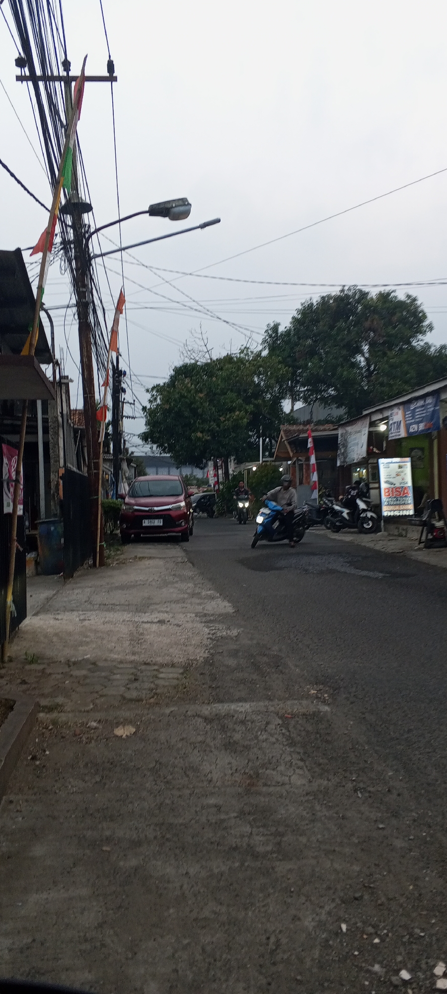 The image captures a street scene in Cibinong, Indonesia, under an overcast sky. A red car with license plate "A 3962 FD" is parked on the left side of the road. Several motorcycles are also present; one is blue and being ridden by a person in a light-colored shirt, another is white and parked further down the road, and a third is black and parked next to the white one. On the right side of the street, there is a building with a sign that says "ATM" and advertisements for "VISA" and money transfer services. A large tree with dark green foliage stands behind the buildings on the right. A pole with numerous electrical wires and three small flags (red and green) is prominent on the left side of the image. The road surface is asphalt, with a paved sidewalk on the left. The overall impression is a moderately busy street in a commercial or residential area.