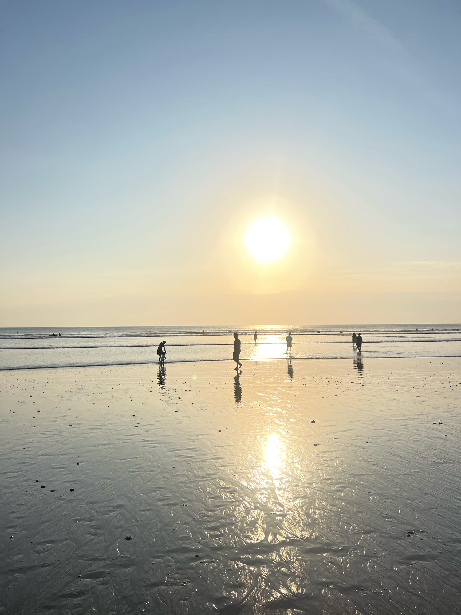 The image captures a serene sunset scene at Kuta Beach, Bali, Indonesia. Several individuals are silhouetted against the bright sun, which is low in the sky and casting a golden reflection on the wet sand and shallow water. The wet sand displays intricate patterns, likely formed by receding waves. In the foreground, the sand is dotted with small pebbles. The horizon is marked by the calm ocean, with gentle waves. The sky transitions from a pale blue at the top to a warm yellow-orange near the sun. The overall atmosphere is tranquil, suggesting a peaceful end to the day.