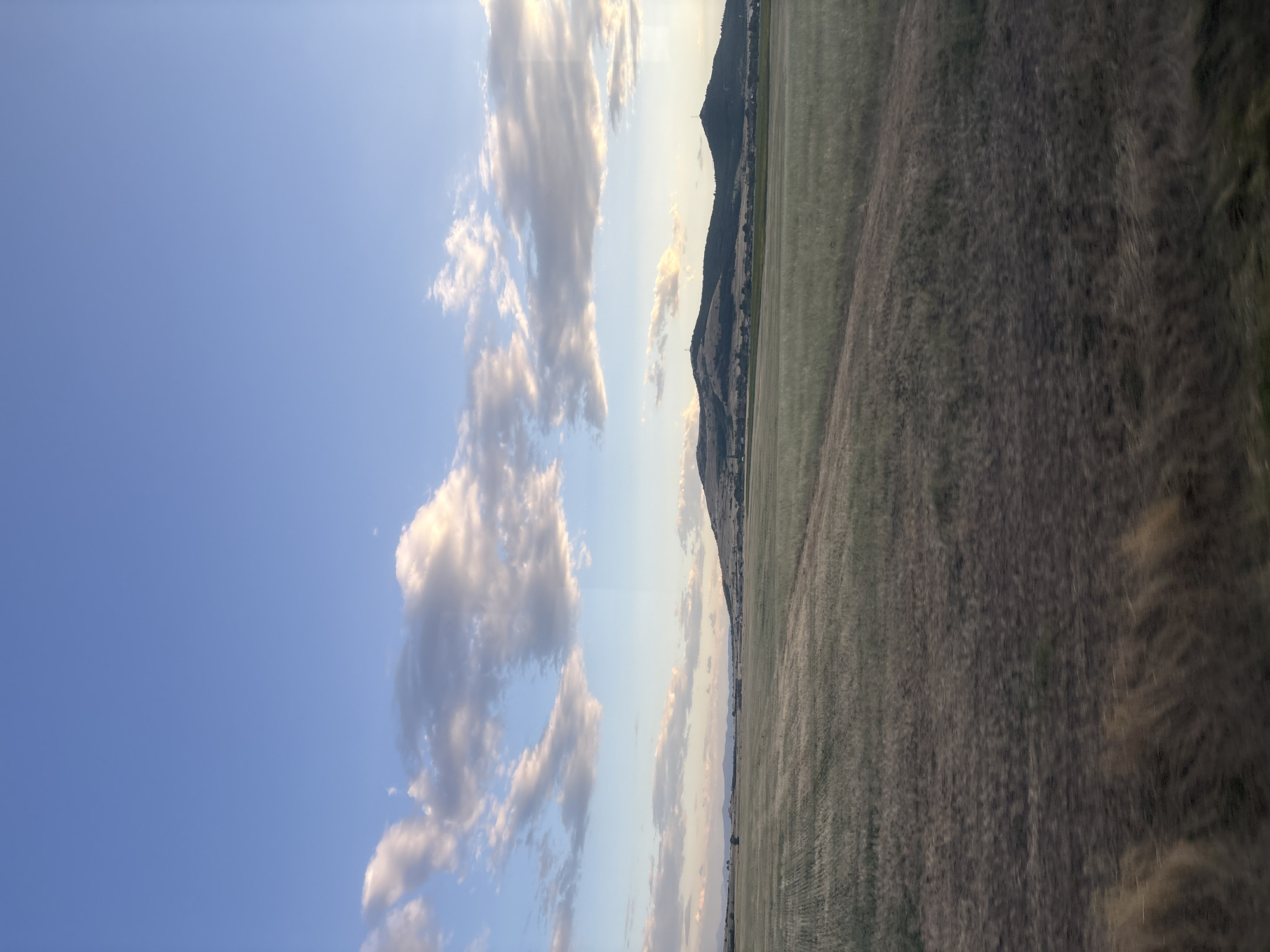 This is a vertical, eye-level, outdoor, landscape photograph taken in the city of Naiden Gerovo, Bulgaria. The scene depicts a rural area under a vast, blue sky filled with scattered, fluffy white and grey clouds. The lower half of the image is dominated by a dry, golden-brown field, suggesting it might be late summer or early autumn. The field appears to have been recently harvested or is fallow, with visible rows and patches of stubble. A dirt path or track runs diagonally across the field, leading towards the background. In the distance, rolling hills covered in dark green vegetation rise from the plain. Several structures, possibly houses or farm buildings, are scattered along the base of the hills, and two prominent cell towers are visible on the hilltops. The overall impression is one of tranquility and open space. There are no people or animals visible in the frame. The lighting suggests it might be late afternoon or early evening, with soft sunlight illuminating the clouds and landscape.