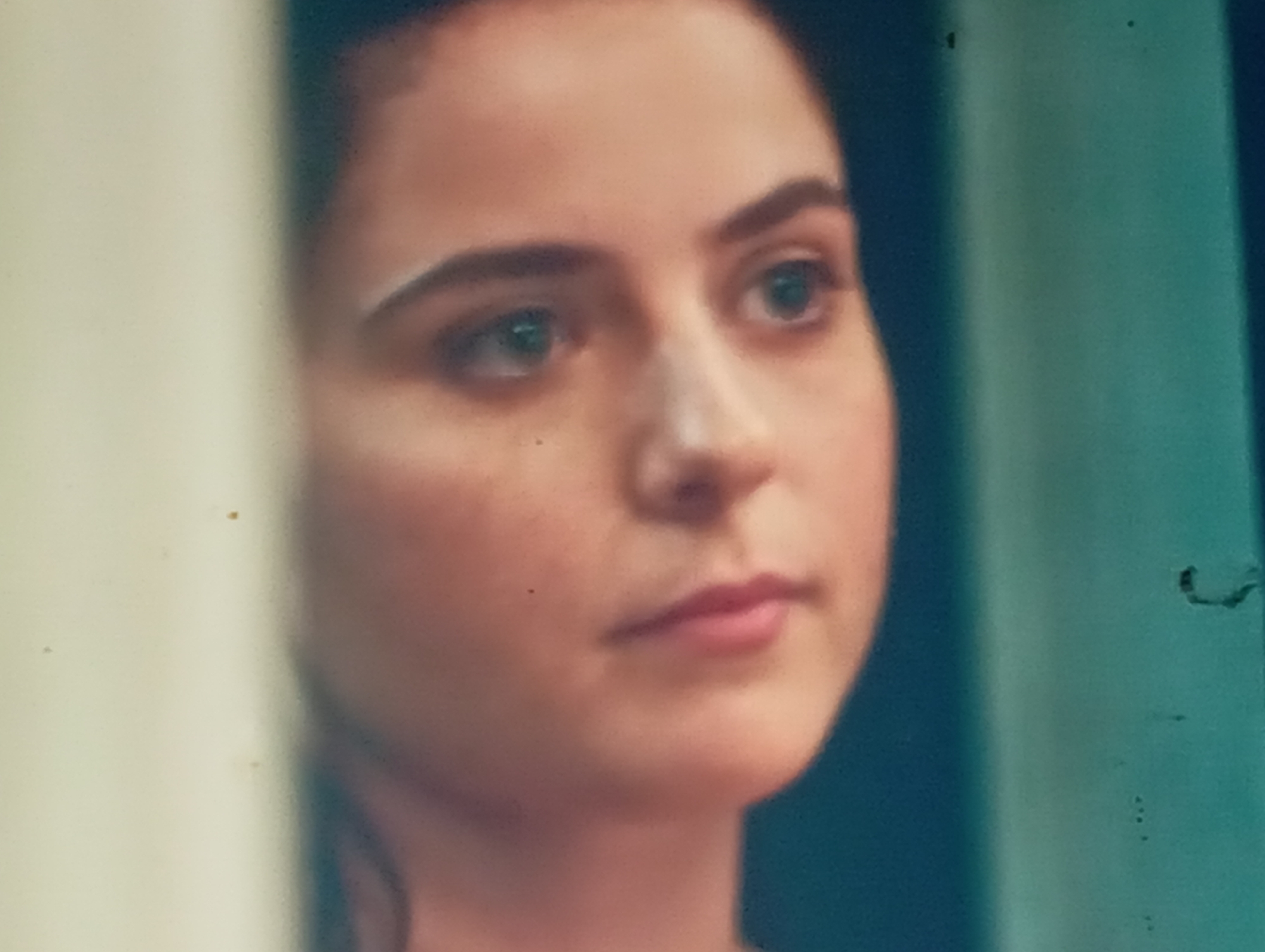 The image is a close-up, slightly blurry, waist-up shot of a young woman with fair skin and dark hair, appearing to be looking to her right. Her eyes are a light blue-green color, and she has a neutral expression. The background is divided into two distinct areas: the left side is a pale, off-white color, possibly fabric or a wall, and the right side is a dark teal, possibly a curtain or a wall. There are some imperfections on the image, including small dark specks that could be dust or damage to the film. The woman appears to be indoors, with soft lighting. There is no identifiable signage or landmarks to pinpoint her location in Sofiivska Borshchahivka, Ukraine.