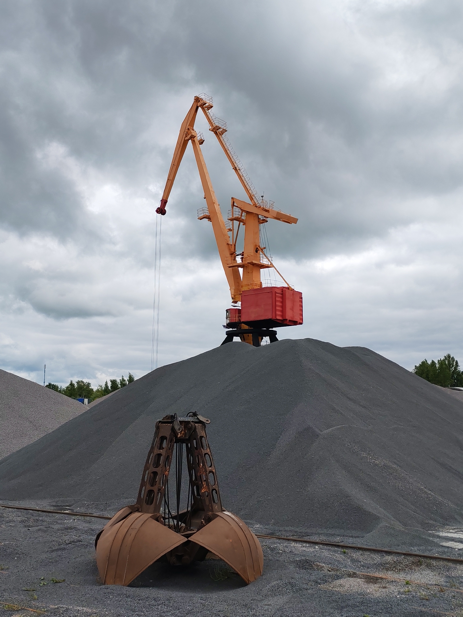 The image shows an industrial scene, likely in Rybinsk, Russia, featuring a large orange port crane standing on a pile of gray gravel or crushed stone. In the foreground, a rusty brown clamshell grab is partially buried in the ground. The sky is overcast with gray clouds. In the background, there are additional piles of similar material and some sparse greenery. No people are visible.