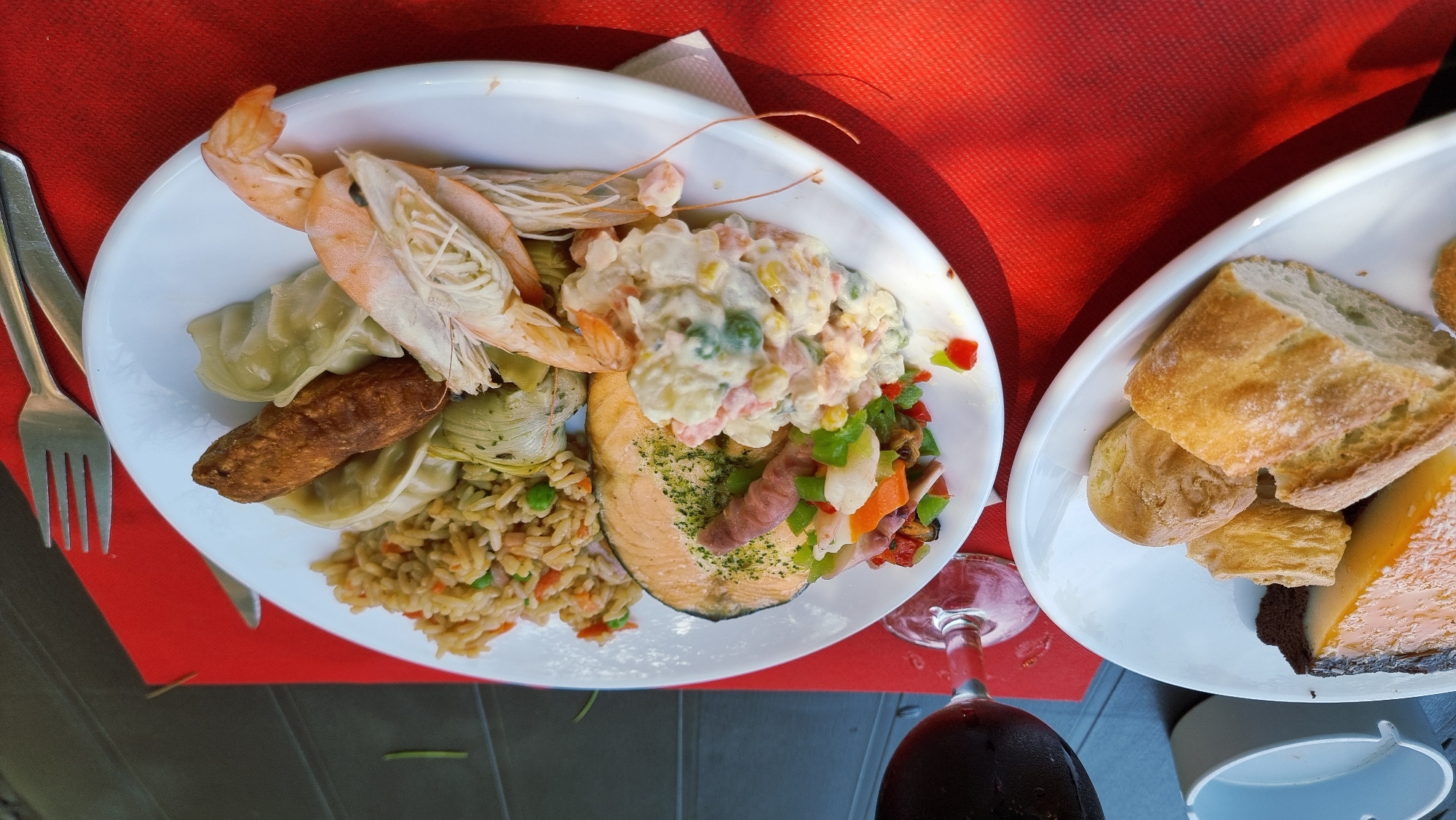 The image shows two white plates of food on a red placemat. The plate on the left contains a slice of cake, some bread rolls, and a slice of bread. The plate on the right contains a slice of salmon, fried rice, dumplings, shrimp, a small salad, and artichoke hearts. A glass of red wine is to the upper left of the plates, and a fork and knife are to the right. The scene appears to be outdoors, possibly at a restaurant in Besalú, Spain, given the typical offerings of such establishments and the rustic wooden table visible in the background.
