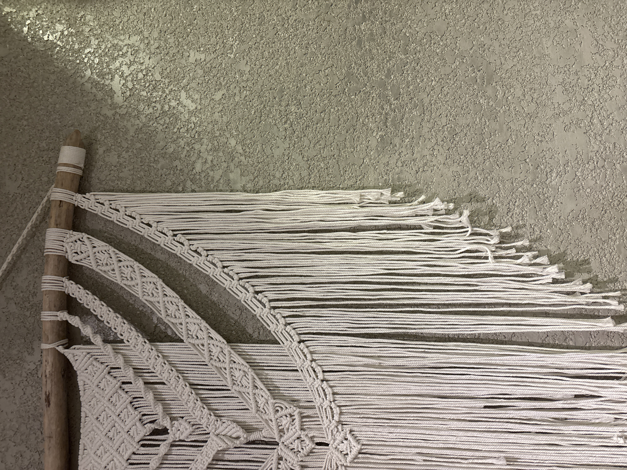 A close-up, low-angle, eye-level shot captures a white macrame wall hanging suspended from a driftwood branch on a textured, light gray wall. The macrame features intricate knotting patterns, forming geometric shapes and a fringe of numerous white cords that hang down. The wall has a rough, stucco-like finish with subtle variations in color and texture, reflecting light and creating shadows. The lighting appears to be ambient, with no direct light source visible. The scene is indoors, against a wall.