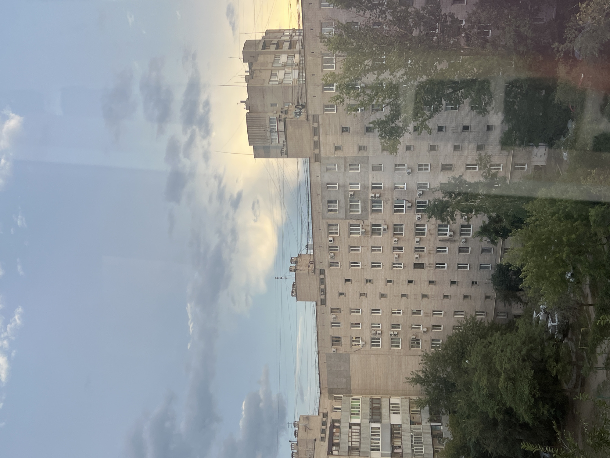 This is an exterior, eye-level, medium shot of apartment buildings in Volgograd, Russia. The buildings are made of concrete blocks and bricks, with numerous windows and balconies. There are trees and greenery in front of the buildings, and the sky is visible above, with clouds and a hint of sunset. The scene appears to be a residential area.