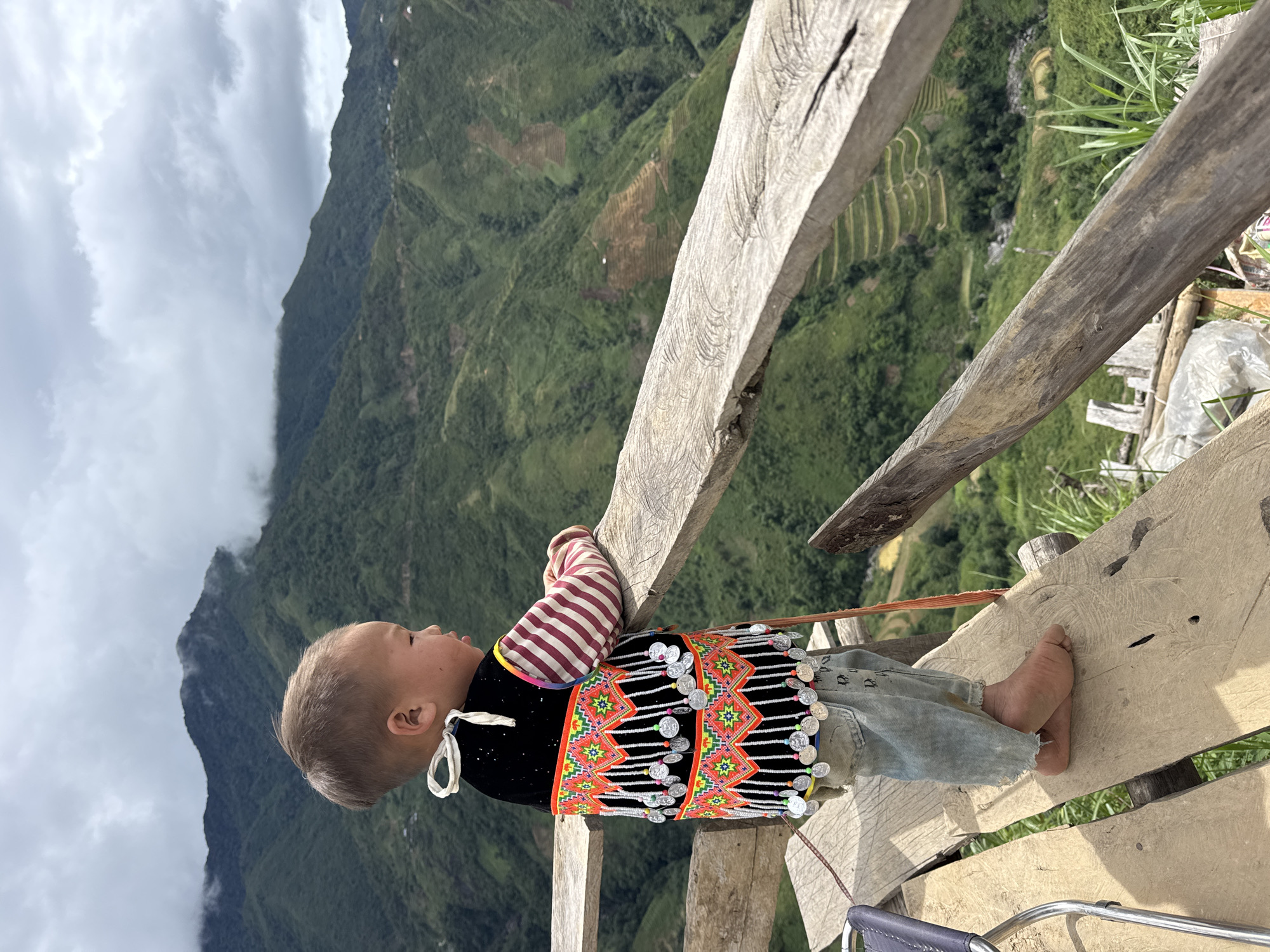 A young child, wearing a traditional vest and striped long-sleeved shirt, stands on a wooden platform overlooking a verdant mountainous landscape in Bắc Yên, Vietnam. The child is facing to the right, looking out at the view. The vest is black with colorful embroidered patterns and adorned with numerous silver coins that hang from strings. The child's light-colored denim pants are torn at the bottom, and they are barefoot. The wooden platform appears to be constructed from rough-hewn planks. In the background, rolling green mountains are visible, with terraced fields carved into the slopes. The sky is overcast with clouds. The scene captures a moment of quiet contemplation for the child amidst a breathtaking natural setting.