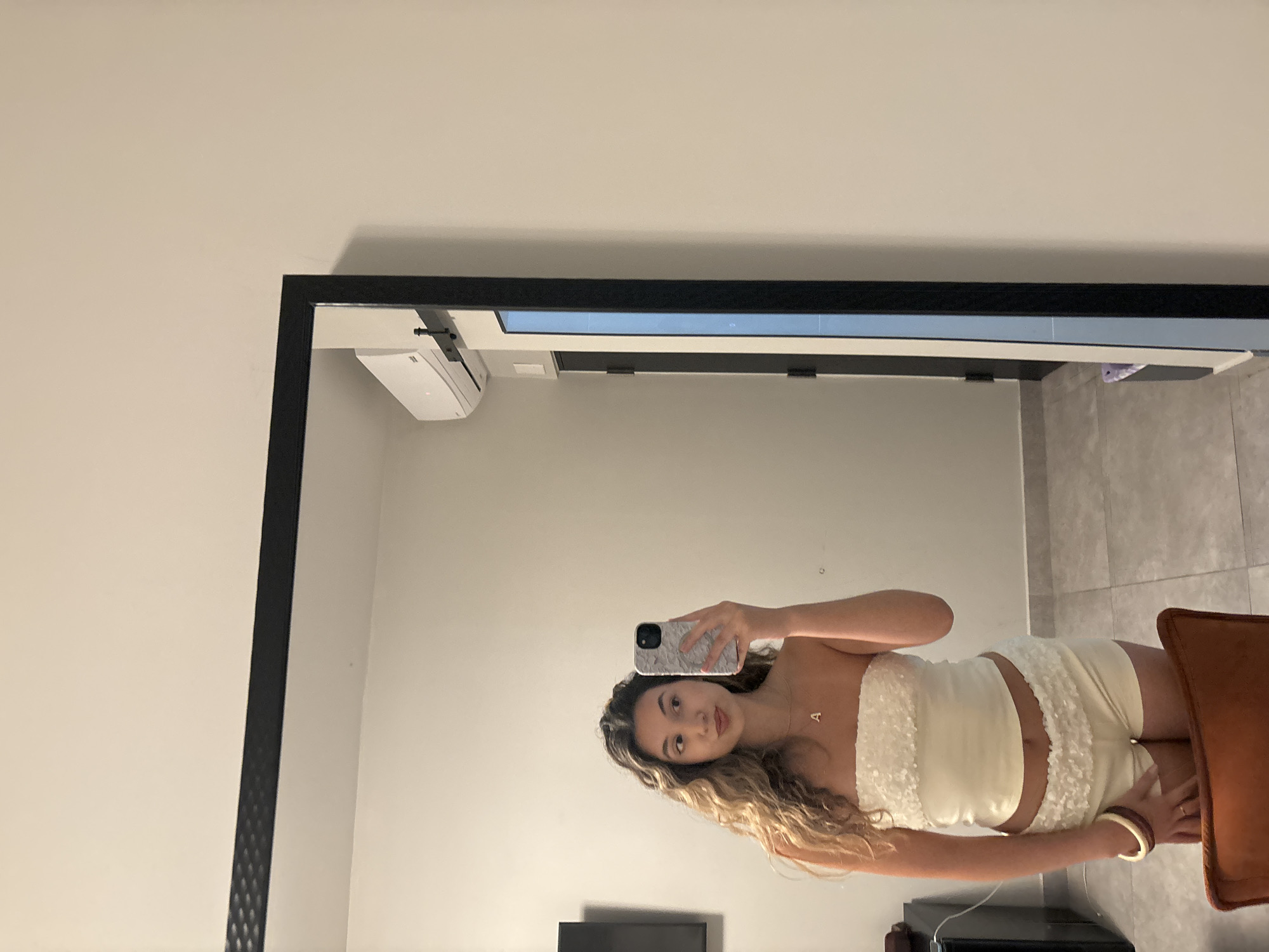 A young woman with long, wavy blonde hair is taking a selfie in a mirror. She is wearing a cream-colored strapless top with ruffled trim and matching ruffled shorts. She has a gold necklace with the letter "A" on it. On her left wrist, she is wearing two bracelets, one brown and one yellow. She is standing in a room with tiled floors and a light-colored wall. A black-framed mirror dominates the left side of the image, reflecting the woman and some of the room. To the right of the mirror, a sliding door with a blue panel is visible. An air conditioning unit is mounted high on the wall above the sliding door. In the background, to the left of the woman, a black flat-screen television is mounted on the wall. The scene appears to be a hotel room in San Juan, Puerto Rico, a territory of the United States.
