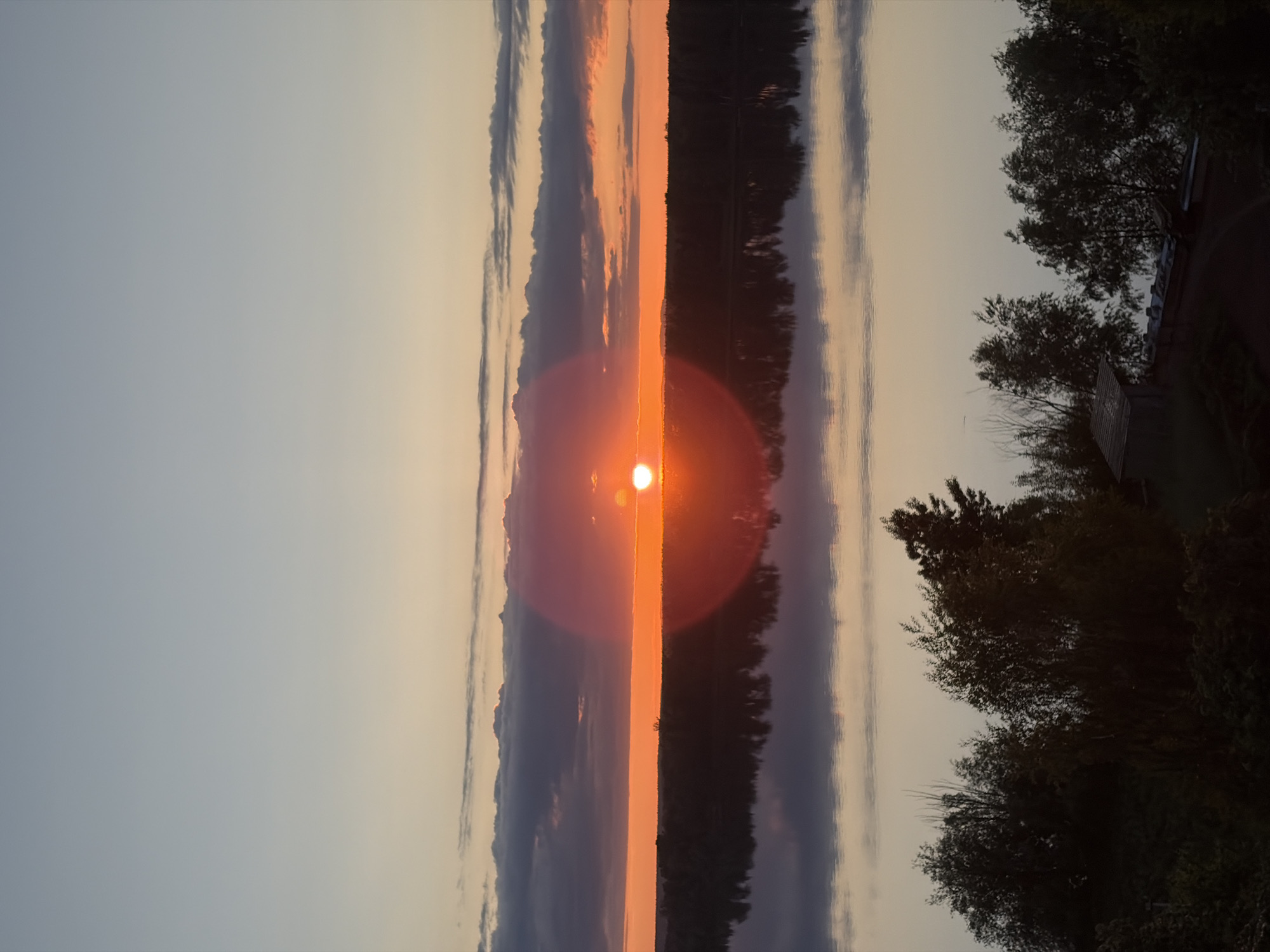 The image depicts a serene sunset or sunrise over a calm body of water, likely a lake or wide river. The photograph appears to have been taken in portrait orientation but is presented rotated 90 degrees counter-clockwise.
If viewed in its correct orientation (rotated 90 degrees clockwise), the scene shows a vibrant sun, intensely orange-red, positioned low on the horizon towards the left side of the frame. A prominent lens flare creates a large, hazy halo around the sun. The sky, occupying the upper portion, transitions from a light blue-grey at the top to warm yellows and oranges near the horizon, with several elongated cloud formations. The calm water, in the lower portion, acts as a perfect mirror, reflecting the sky, clouds, and a brilliant, vertical column of orange and red light extending directly from the sun downwards.
On the right side of the image, a dark, silhouetted shoreline is visible, densely covered with trees and foliage, whose forms are also reflected in the still water. A small, indistinct structure, possibly a building or dock, is partially obscured by the trees. The low sun and intense warm colors unequivocally indicate either sunrise or sunset. The scene evokes a feeling of peace, serenity, and natural beauty. There are no people or visible text in the image.