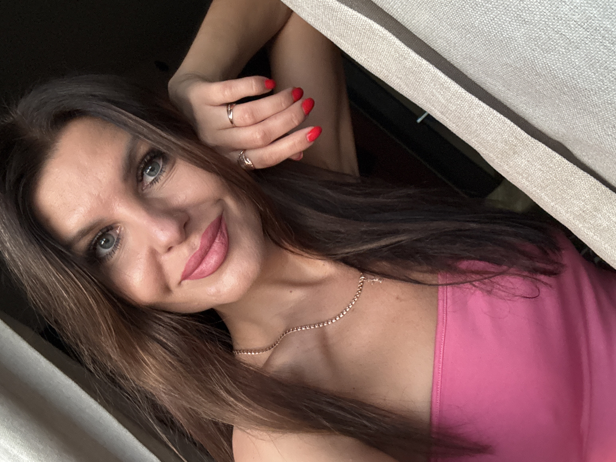 This media file is a close-up, selfie-style portrait of a woman with long, brown hair and striking blue eyes. She is looking directly at the camera with a soft smile. Her lips are painted a vibrant pink, and she wears makeup that accentuates her features. She has bright red nail polish on her fingers and is wearing two delicate rings on her left hand. A thin, rose-gold necklace with small, clear stones adorns her neck. She is wearing a bright pink, sleeveless top. The background is dimly lit, with a textured, light-colored fabric partially obscuring the right side of the image, suggesting she is near a window with curtains. The lighting indicates it is likely daytime, possibly late afternoon given the soft shadows. The overall mood is relaxed and confident. There is no visible text in the image. The location context provided is Chaikovsky, Russia.