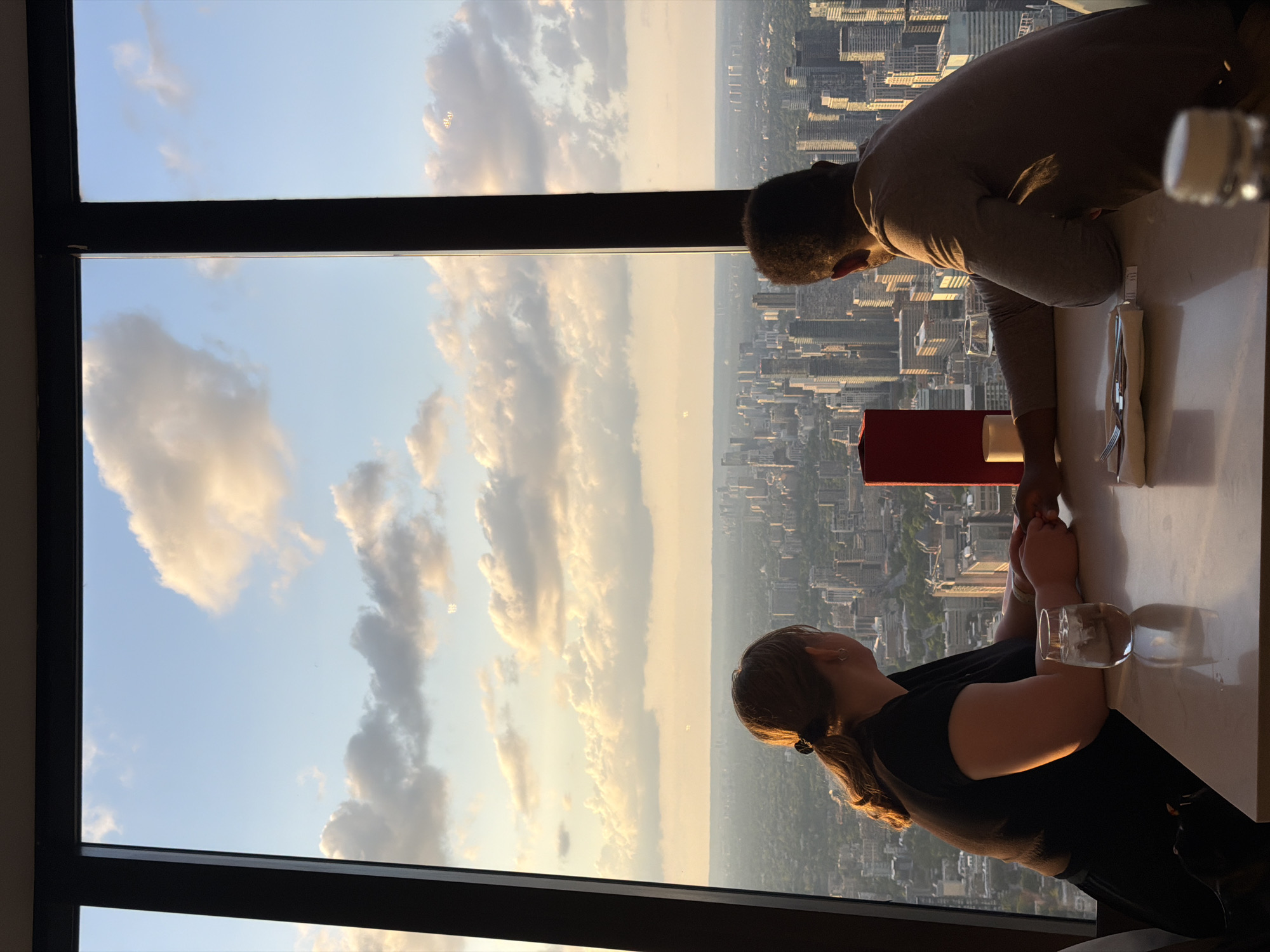 A high-angle, eye-level photograph captures an interior restaurant scene looking out onto a sprawling cityscape under a cloudy, golden-hour sky. Two individuals are seated opposite each other at a light-colored table, facing a large window that frames the panoramic view.
The individual on the left is a female with light brown hair tied in a ponytail, wearing a black short-sleeved top. Her back is mostly to the viewer, but her left profile is visible as she gazes out at the city. A small, round earring is visible on her left ear. Her left arm rests on the table, and her right hand is clasped with the left hand of the individual opposite her.
The individual on the right is a male with short, dark, curly hair, wearing a light brown or beige long-sleeved shirt. His back is mostly to the viewer, with his right profile visible as he also looks out at the city. His right hand is clasped with the female's right hand across the table.
On the table in front of the female, a clear glass filled with ice and water reflects the warm ambient light. Centered on the table is a tall, slender red object, possibly a menu holder or a decorative item, with a smaller white object (likely a candle or cup) beside it. In front of the male, a neatly folded white napkin holds a fork and knife. A blurred white bottle cap is visible in the extreme foreground on the right.
Outside the window, a vast urban landscape of Toronto unfolds, characterized by numerous high-rise buildings and skyscrapers extending to a hazy horizon. The sky above is light blue with scattered cumulus clouds, illuminated by the warm, golden light of the setting sun, creating a soft glow on the cloud edges. Distinctive snowflake-like light reflections, consistent with the anti-glare coating of the 360 Restaurant in the CN Tower, are visible on the window panes. The interior of the restaurant is dimly lit compared to the bright exterior, emphasizing the view.