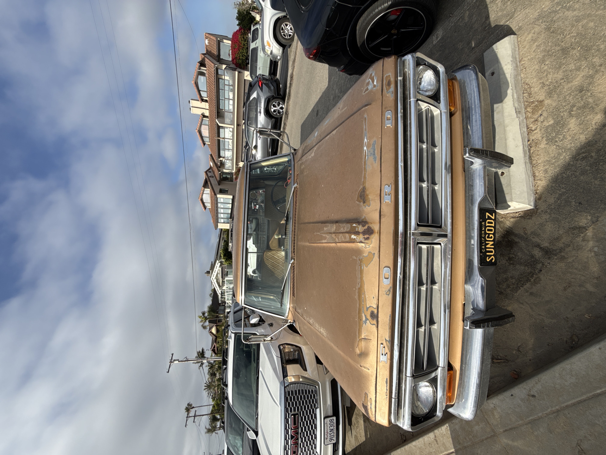A street scene in Encinitas, United States of America, is captured on a partly cloudy day. In the foreground, a vintage, light brown Ford pickup truck is prominently displayed. Its hood shows significant wear with peeling paint and rust, and the "FORD" emblem is debossed near the front. The truck features a chrome grille, round headlights, and a shiny chrome front bumper. A black California license plate with yellow text, reading "SUNGODZ," is attached to the bumper.
Behind the Ford, a modern white GMC pickup truck is partially visible, showcasing its distinctive grille with the "GMC" logo and a California license plate with the number "9USN308". Further down the street, other vehicles, including a dark SUV with black multi-spoke wheels and a grey SUV, are parked.
The background reveals a residential setting with several multi-story houses featuring brown tiled roofs and numerous windows, likely overlooking the street. Palm trees are interspersed among the houses, contributing to the local flora. Power lines stretch across the upper portion of the frame against a sky that is a mix of blue patches and white clouds. The vehicles are parked on pavement or a street surface visible in the lower and right sections of the image.