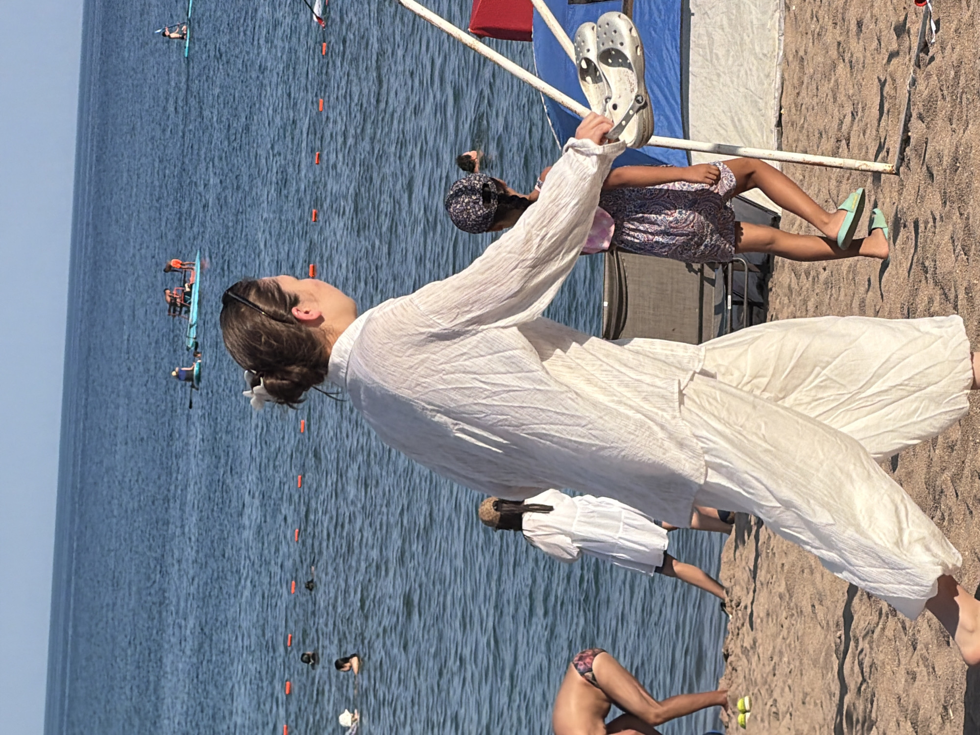 In a beach scene, a woman in a white linen outfit walks on sand, holding a pair of white clogs. She is in the foreground, facing away from the camera. Behind her, a young girl in a patterned dress and a floral cap walks towards a blue and red tent. In the background, several people are in the water, some swimming and others on paddle boats. The water is calm, and the sky is clear and blue. The environment appears to be a public beach with a body of water and sandy shoreline.