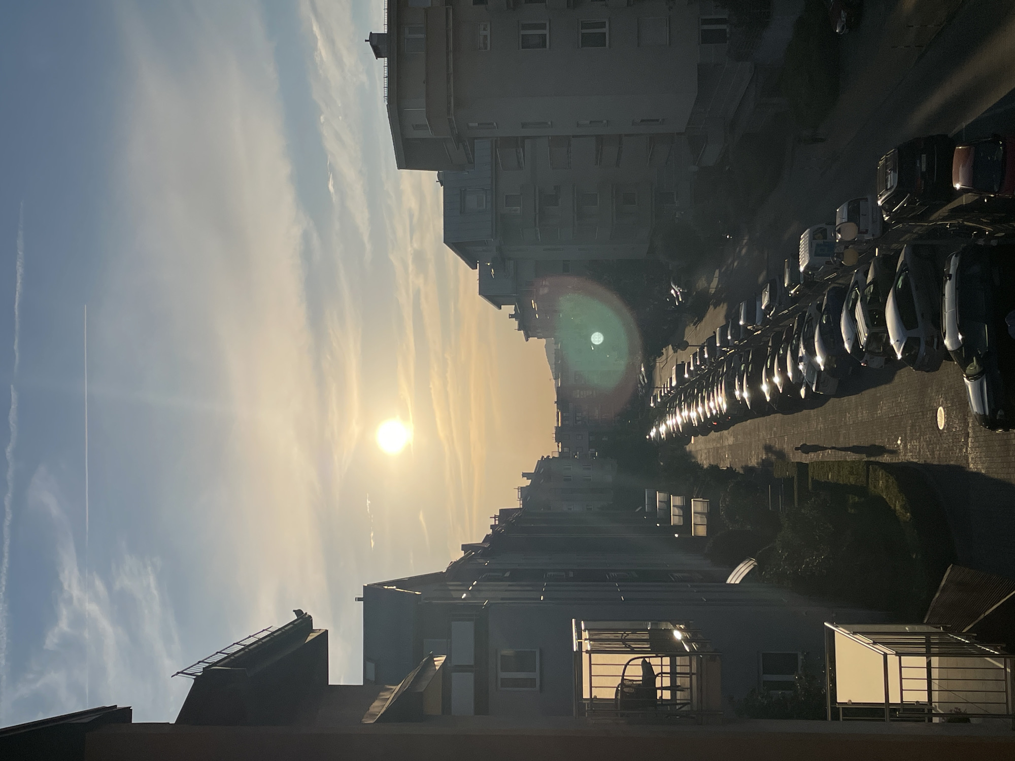 The image depicts a street scene at dusk, likely in an urban environment. The sun is low on the horizon, casting a warm, golden light across the sky and creating long shadows. A row of parked cars lines the street, their headlights reflecting the ambient light. Buildings flank the street, appearing to be multi-story residential or commercial structures. The sky is a mix of blue and orange, with wispy clouds. One building has a notable lens flare from the sun. A single person is visible walking on the pavement, casting a long shadow. The overall mood is serene and calm, characteristic of the end of the day. The image appears to be taken from a higher vantage point, possibly a window or balcony. There is no visible text.