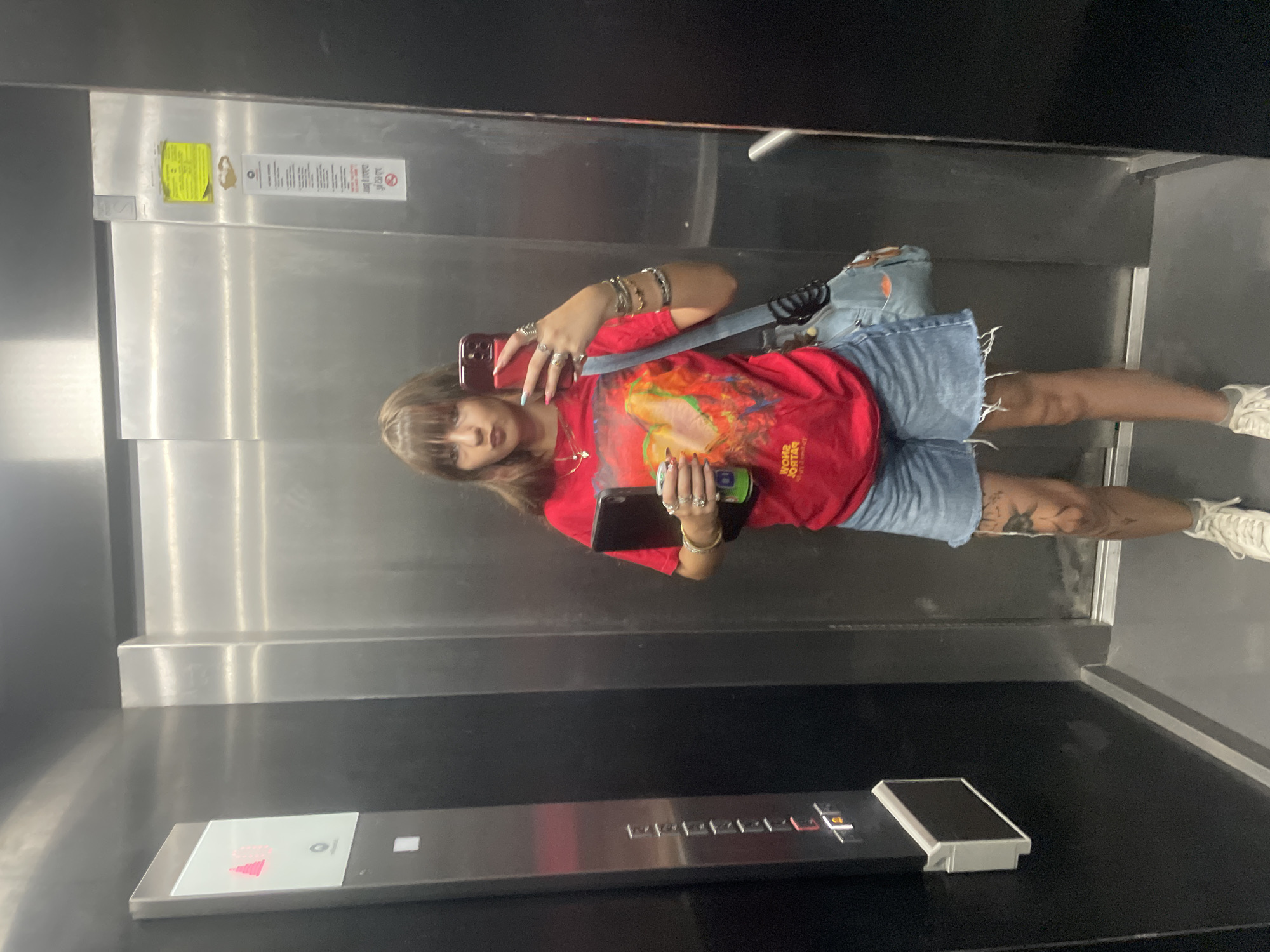 The image depicts a person standing inside an elevator. The person is wearing a red t-shirt with a graphic print, denim shorts, and white sneakers. They are holding a smartphone in their right hand and a green can in their left. The elevator interior is mostly made of stainless steel, with some black paneling at the bottom. A control panel with buttons and a digital display is visible. The image is taken from a low angle, with the camera tilted. The lighting appears to be artificial, consistent with an indoor setting. The person's expression is neutral. There is some text on a yellow sticker on the elevator wall, which is partially visible and unreadable. The scene is likely taking place during the daytime, as indicated by the general brightness of the environment, though specific time of day cannot be determined. The location is identified as Raanana, Israel.