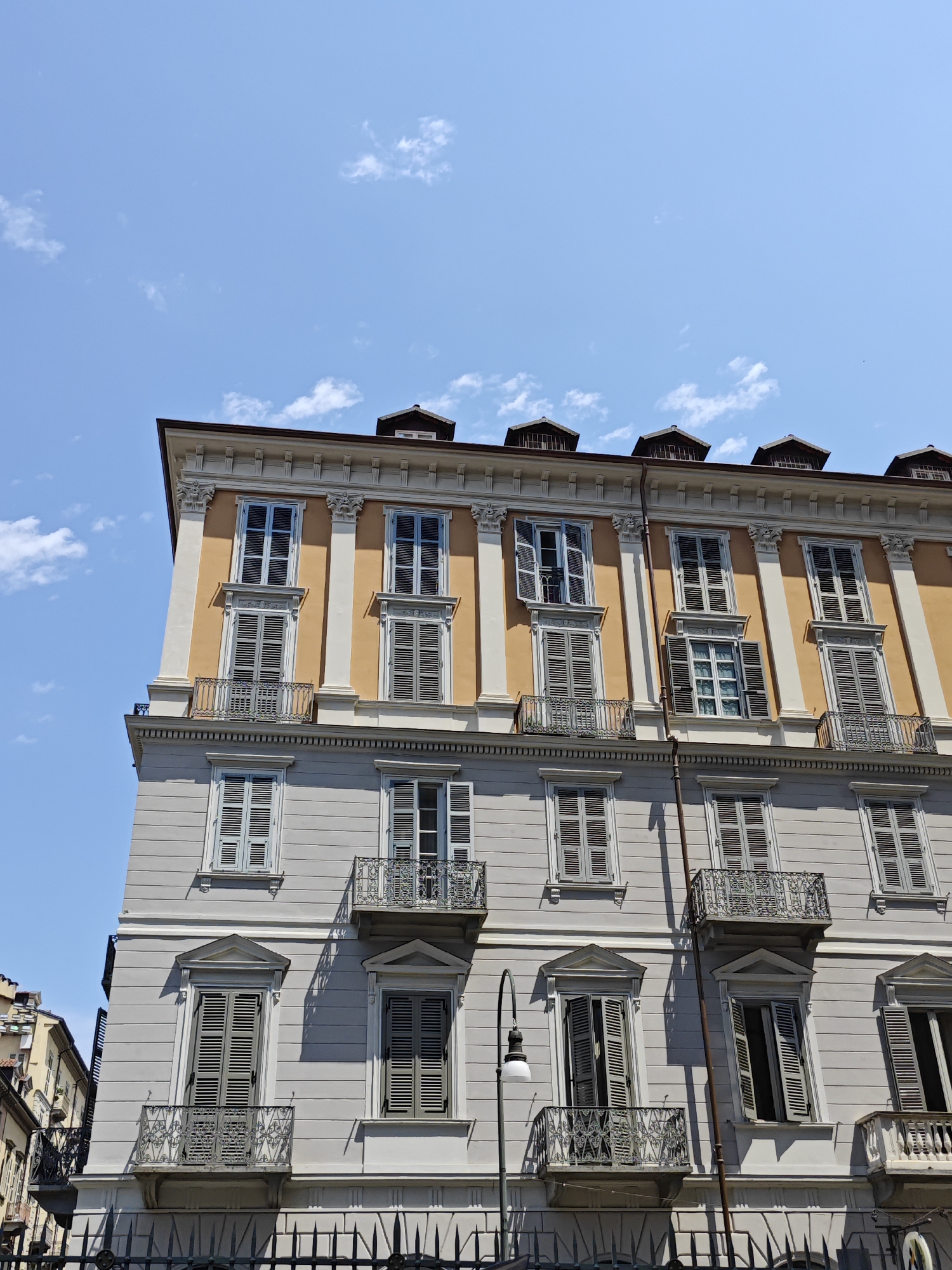 This is a low-angle, full shot of a building facade in Turin, Italy, on a bright, sunny day with a clear blue sky and scattered white clouds. The building is multi-story, with ornate architectural details. The upper story is painted a light orange-yellow, while the lower stories are a light gray. Both stories feature numerous windows with closed wooden shutters. Some windows have decorative balconies with wrought-iron railings. A dark metal fence is visible at the bottom of the frame. A streetlamp with a white glass shade is attached to a pole that runs vertically along the building. The building is designed in a classical style, with pilasters and cornices. The image captures the historic architecture of the city.