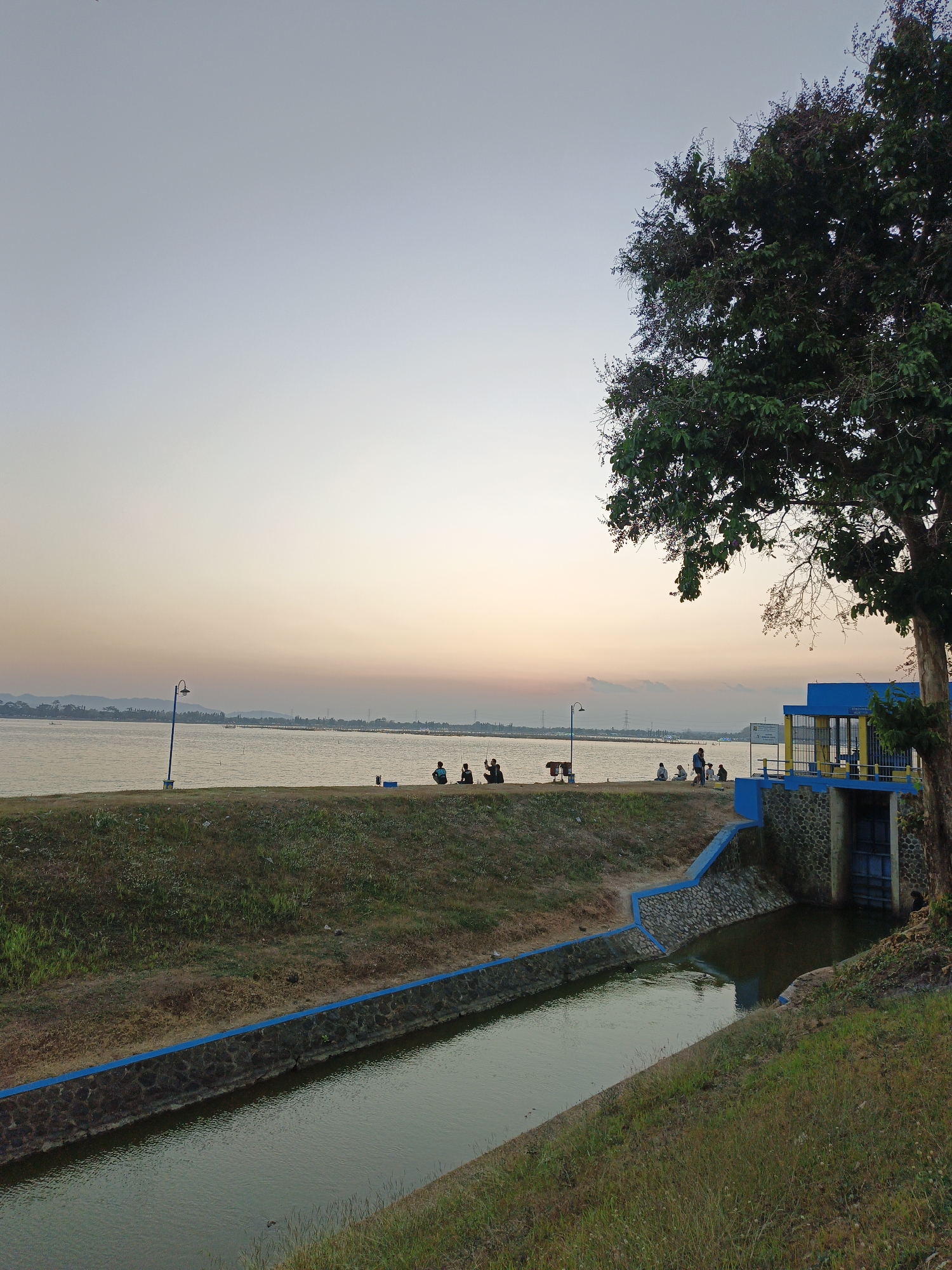 The image depicts a serene waterscape at dusk, likely in Krakitan, Indonesia, based on context. A wide body of water, possibly a reservoir or lake, stretches across the horizon, with low-lying hills visible in the distance. The sky transitions from a pale blue overhead to a warm gradient of orange and yellow near the horizon, suggesting sunset.
In the foreground, a man-made canal with stone-lined banks and a blue trim guides water away from the larger body. The canal leads towards a structure that appears to be a water management facility, featuring a blue building with yellow accents, stone foundations, and visible gates. A large, leafy tree with dark green foliage dominates the right side of the frame, its branches reaching towards the sky.
Along the embankment overlooking the water, several individuals are scattered. Most are seated or crouched, some appearing to be fishing, indicated by the presence of fishing rods. A lamppost with a blue pole stands near the water's edge. The overall environment is grassy and somewhat overgrown in places, with a sense of natural beauty meeting human infrastructure.
