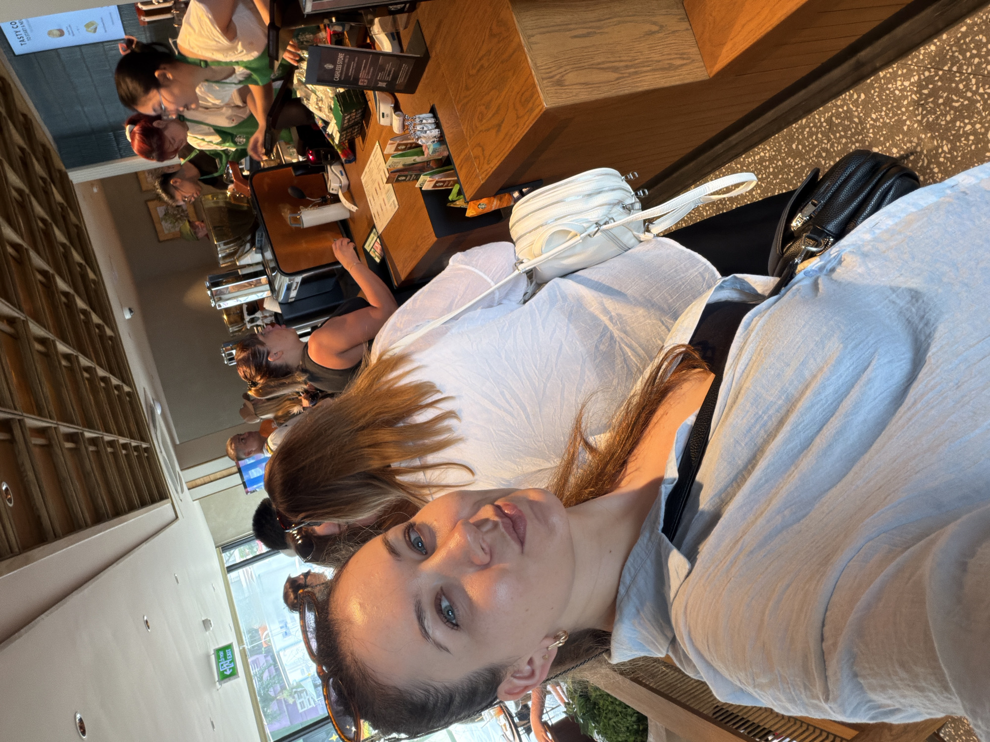 The image depicts the interior of a Starbucks coffee shop in Đà Nẵng, Vietnam, likely located in a mall or a busy urban area, as indicated by the visible "EXIT" sign and the architectural style suggesting a modern commercial establishment. The foreground is dominated by a woman taking a selfie, her face angled towards the camera with blue eyes and light brown hair. She is wearing a light blue collared shirt and a black top underneath, with a black crossbody bag slung over her shoulder and a white handbag hanging from it.
Behind her, the bustling atmosphere of the coffee shop is evident. Several other patrons are visible, some in line and others interacting with baristas at the counter. The baristas, identifiable by their green Starbucks aprons, are engaged in serving customers and preparing drinks. One barista with red hair and glasses is seen interacting with a customer at the register. Another barista, wearing glasses and a black shirt under her apron, is further down the counter.
The counter area is well-stocked with various items, including coffee packets, cups, and POS systems. A "CASHLESS STORE" sign is prominently displayed, indicating a payment preference. The background reveals the characteristic Starbucks interior design with wooden ceiling panels and warm lighting. Through the large windows behind the counter, a glimpse of the city outside is visible, with trees and buildings suggesting an urban environment. The floor is a speckled pattern. The overall scene captures a typical day at a popular coffee chain, with a focus on customer service and a lively ambiance.