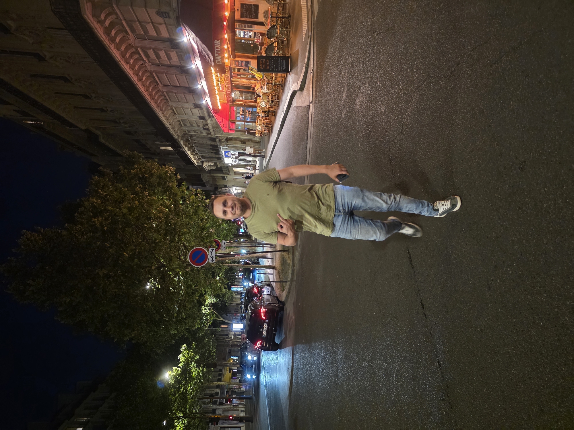 A man with a bald head and a short beard walks down a street at night in Paris, France. He is wearing a green t-shirt and blue jeans, and he is pointing his left index finger towards the camera. The street is wet, suggesting recent rain. In the background, there are cars driving on the street, and a building with a red awning that appears to be a restaurant. Trees line the sidewalk, and streetlights illuminate the scene. The image was taken at street level.