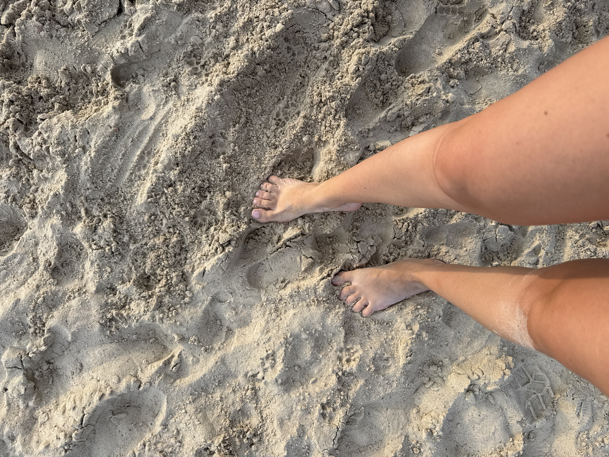 The image displays a close-up, overhead view of a person's legs and feet standing on sand. The sand is light brown and appears textured with footprints and rippled patterns. The person's legs are tanned, and their feet are bare, with toes visible. The lighting suggests it is daytime, possibly late afternoon, given the shadows cast on the sand. The location is identified as Karachunivka, Ukraine.