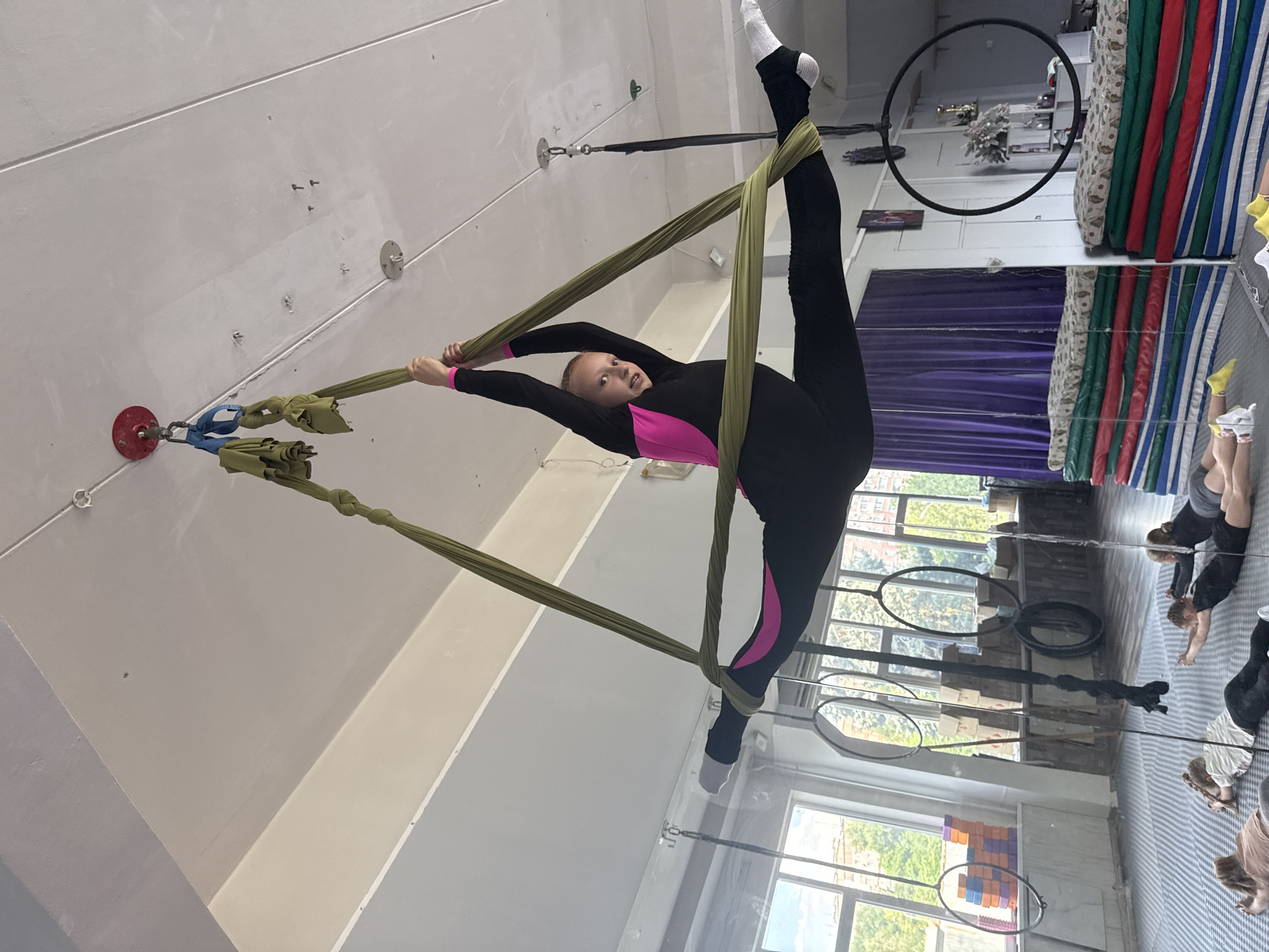 The media file captures an indoor scene, likely a gymnastics or aerial arts studio in Dnipro, Ukraine. The primary subject is a young female aerialist performing an acrobatic pose on a piece of aerial fabric. She is dressed in a black full-body suit with bright pink accents on the sides of her legs and across the chest. Her legs are split in a vertical split, with one leg extended straight up and the other pointing down. The aerial fabric, a pale green color, is wrapped around her legs and hands, supporting her in the air. Her expression is focused and determined.
The setting features a high ceiling with visible lighting fixtures and mounting points for aerial equipment. A large mirror covers a portion of the wall, reflecting other individuals in the room, some of whom are also engaged in exercises on the floor. In the background, stacks of colorful mats are visible, along with large windows that offer a glimpse of the outdoors, suggesting it's daytime. The overall atmosphere appears to be one of training and athletic pursuit. The orientation of the image suggests it was taken with the camera held vertically.