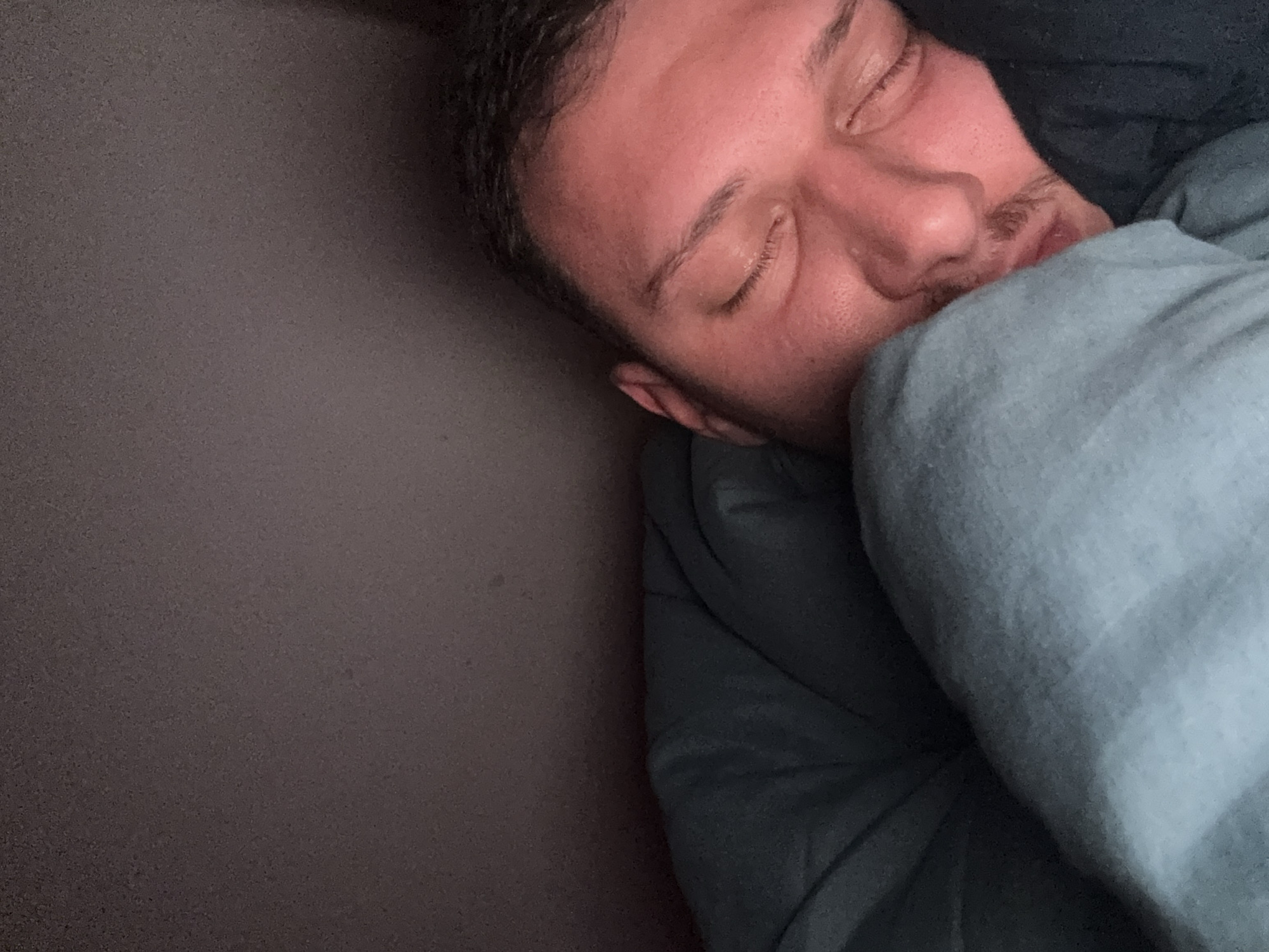 A man is asleep in bed. He is on his right side and covered with a blue blanket. He is lying on a gray surface, possibly a mattress. The location is unknown and cannot be determined from the image.