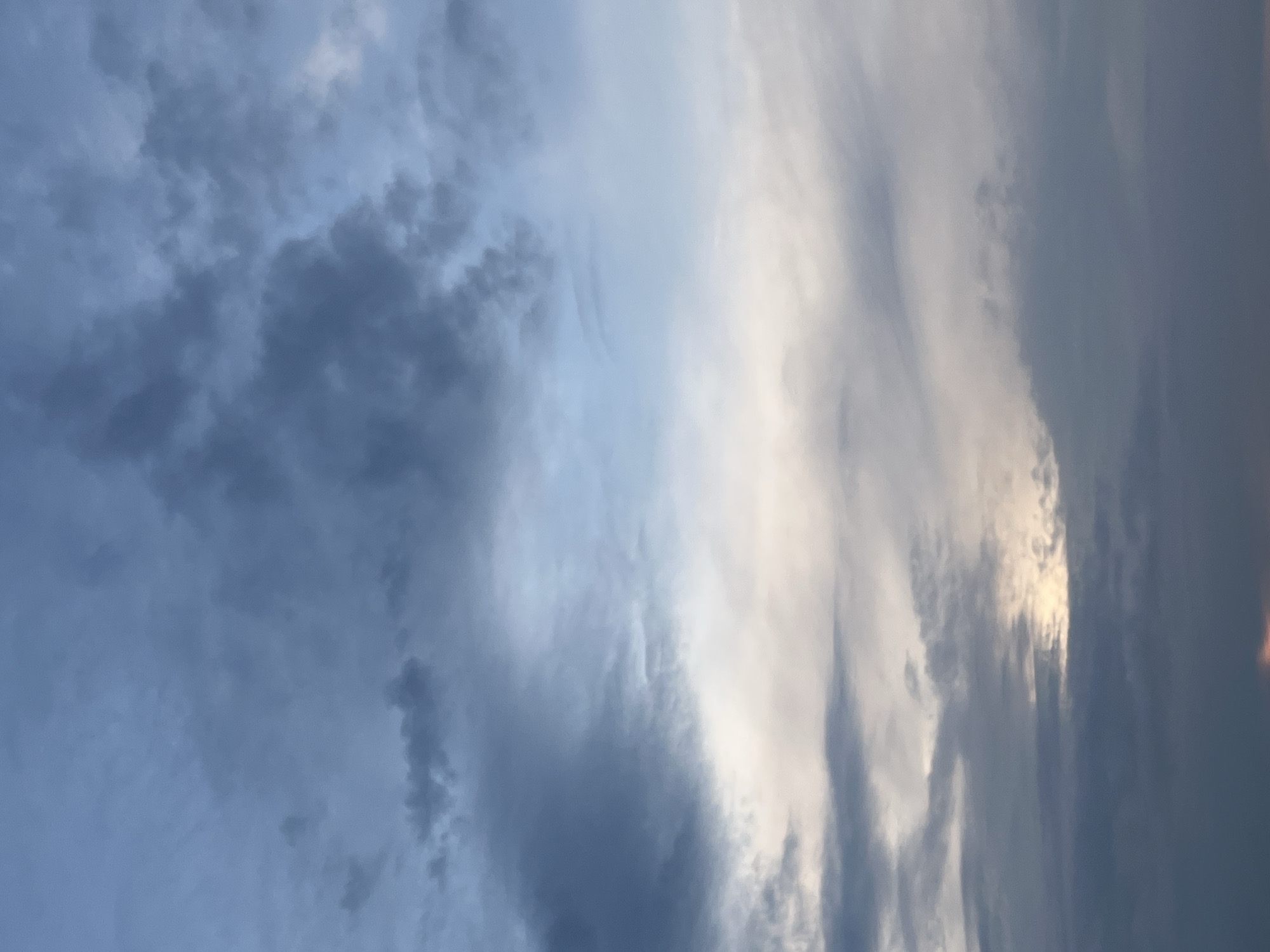 The image shows a cloudy sky, with varying shades of gray and blue. There are darker, dense cloud formations on the left side, contrasted by lighter, wispy clouds towards the right. The lighting suggests that the sun may be setting or rising, with some clouds appearing brighter due to the light. There is no other information available to confirm the exact location, time, objects, people, and action.