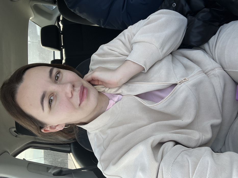 A single female subject is depicted inside a vehicle, at an unspecified location within the city of Shchepkin, Russia. She is positioned in what appears to be the front passenger seat, facing forward and looking directly at the camera with a slight smile. The woman has brown hair, visible eyes, and light skin, wearing small silver hoop earrings and a delicate silver necklace. She is dressed in a light beige, ribbed bomber-style jacket, partially unzipped, revealing a light pink shirt underneath. Her left hand is raised, with her fingers lightly touching her neck/chin area.
The vehicle's interior features black upholstered seats and headrests in the background. To the subject's right, a portion of another individual's arm or shoulder, clad in a dark blue or black puffy jacket, is visible. Through the vehicle's side window on the left, an outdoor scene is visible, featuring a light-colored vertical structure, possibly a fence or building wall, under natural daylight. A small purple object is partially visible at the bottom center of the frame.