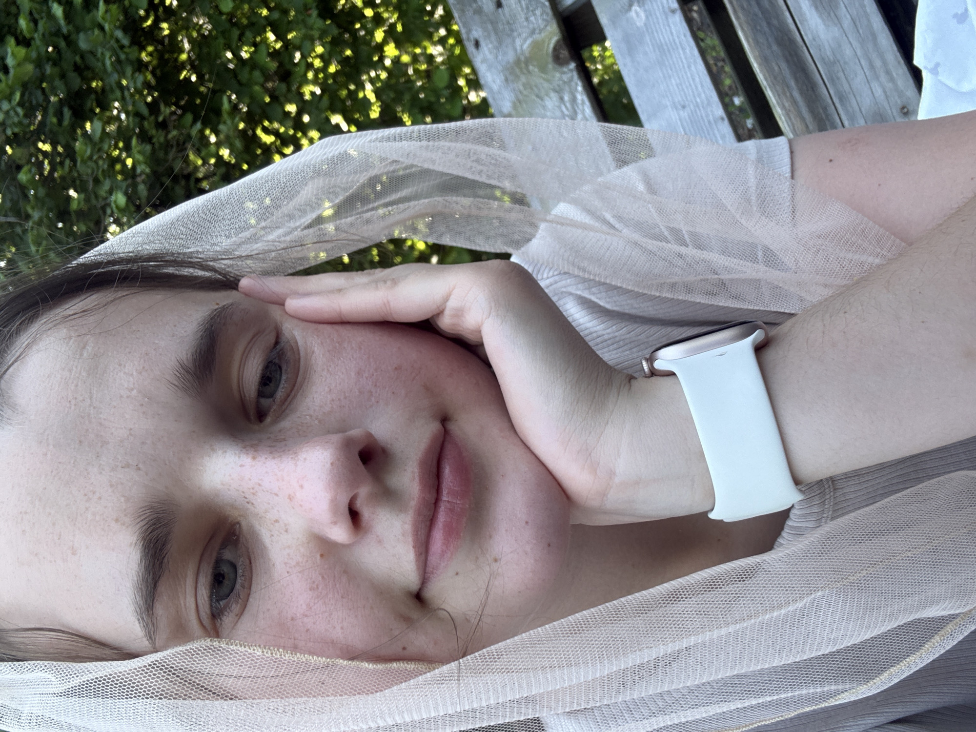 The image is a close-up selfie of a young person with fair skin, freckles, and light blue eyes. They are resting their chin on their hand, with a light-colored sheer veil covering their head and shoulders. The veil has a subtle shimmer. The person is wearing a light-colored ribbed top and a white smartwatch on their left wrist. The background consists of blurred green foliage and a wooden bench, suggesting an outdoor setting. The scene does not contain information pertaining to Beltershain, Germany.