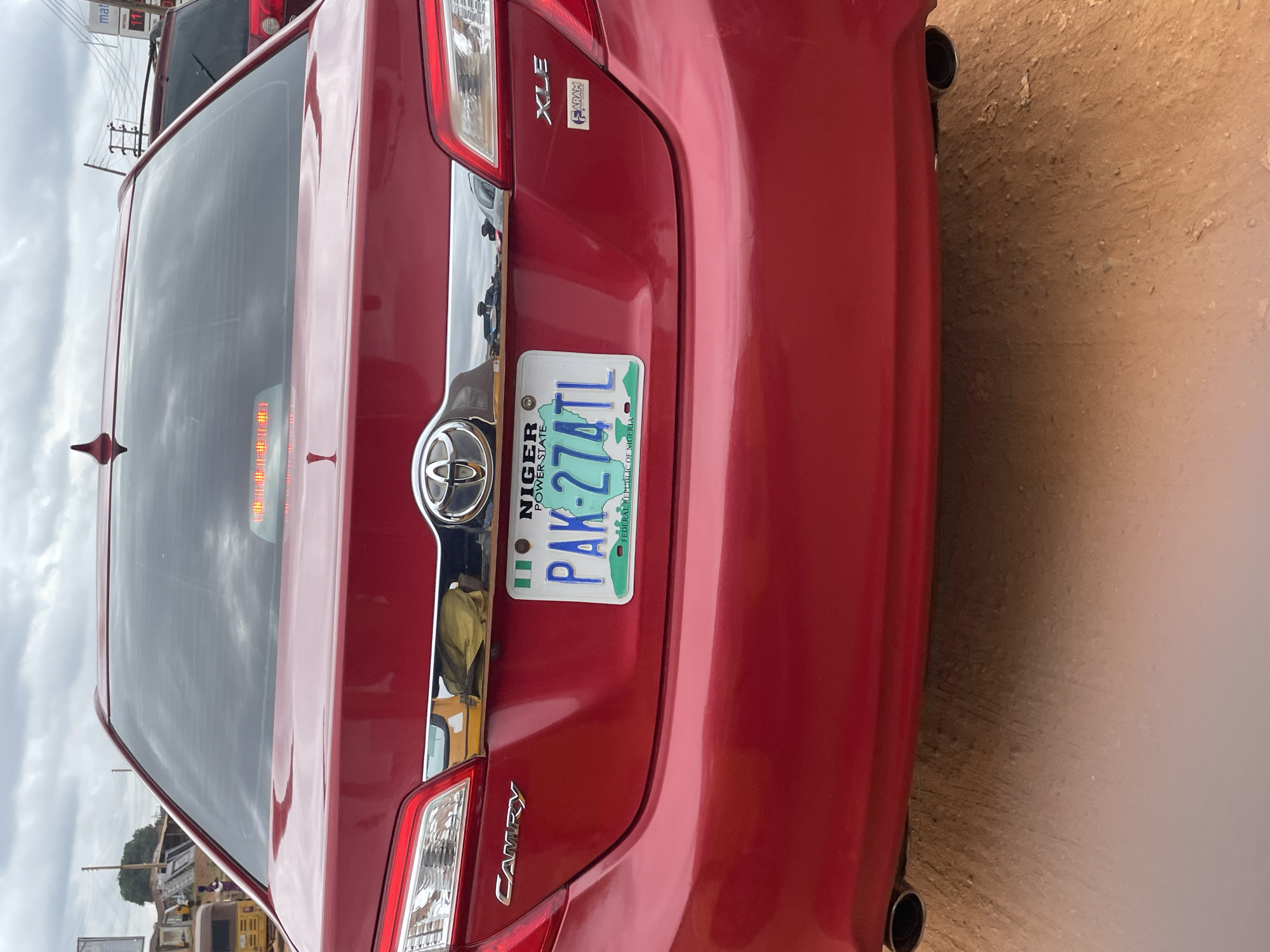 The rear view of a red Toyota Camry XLE sedan is shown. The vehicle is parked on a dirt road. The license plate reads "PAK-274TL" and is from Niger State, Federal Republic of Nigeria. The car has dual exhaust pipes. In the background, there are some low-rise buildings and utility poles, suggesting an urban or semi-urban environment within Minna, Nigeria. The sky is overcast.