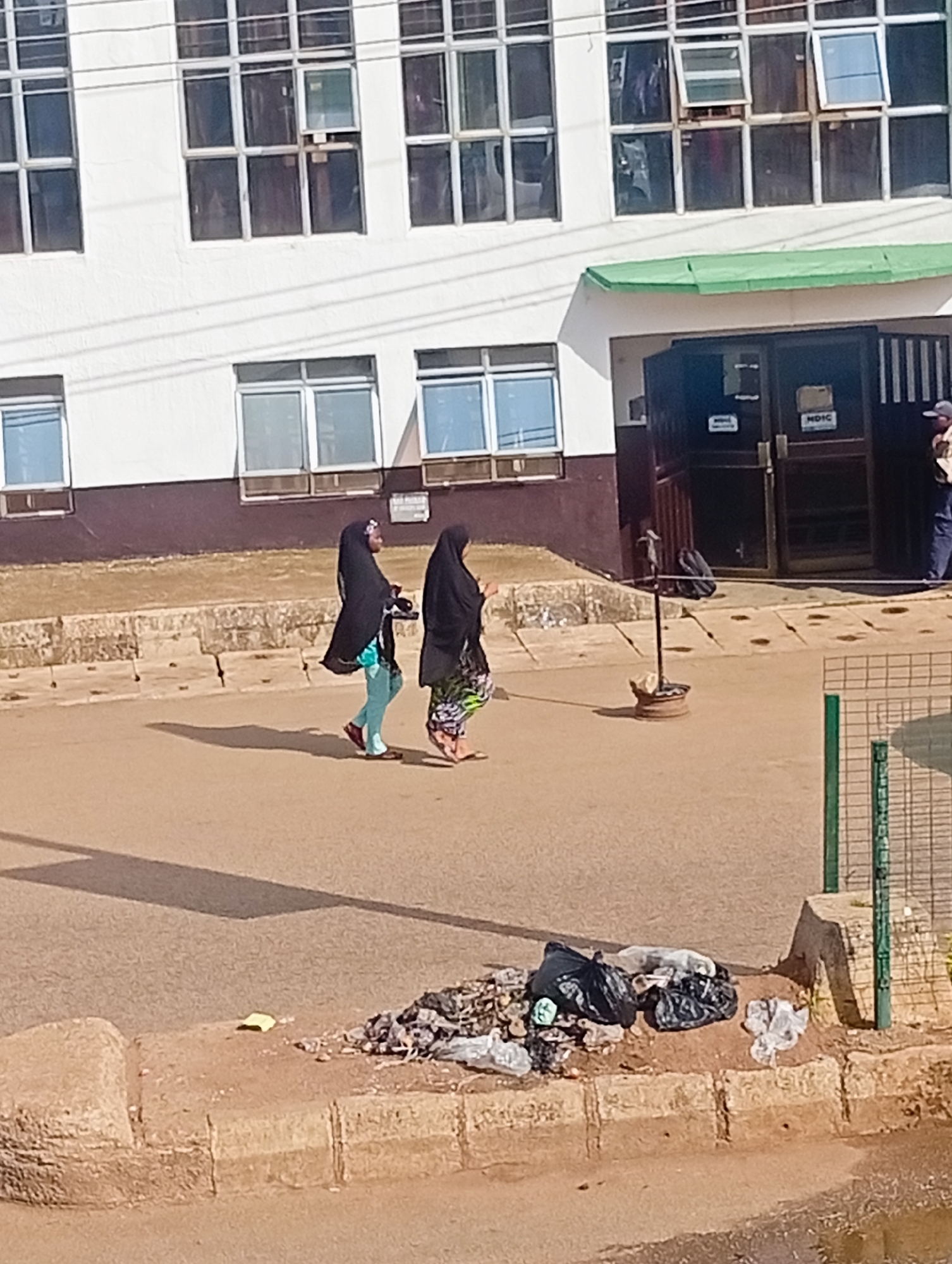The image depicts a street scene in Bukuru, Nigeria. In the foreground, a pile of refuse, including black plastic bags and other debris, sits on a dirt patch next to a concrete curb. The street surface is a light brown asphalt. Further back, two women are walking on the street, both wearing hijabs. One woman is wearing a black hijab and a light blue garment, while the other wears a black hijab and a colorful patterned skirt. They are casting shadows on the asphalt. Behind them, a building with multiple windows is visible, with some windows open. A green awning extends over the entrance to the building, which features double doors and a metal fence. A person wearing a cap and a light-colored shirt is standing near the entrance. A pole with a circular base and what appears to be a rock or small item on top stands on the pavement. A wire mesh fence with green posts is also present on the right side of the image.