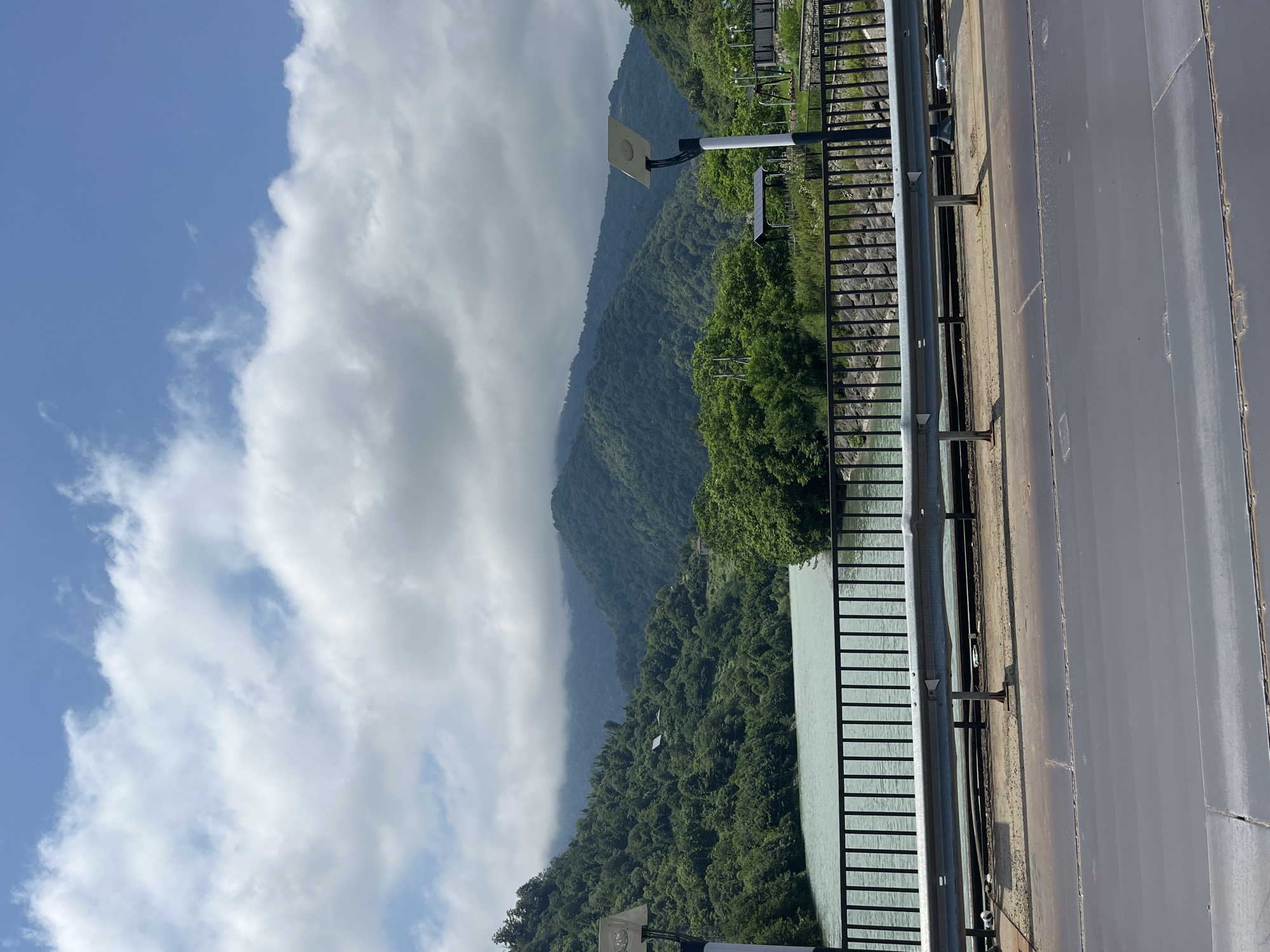 The image shows a scenic view of the Adjaristskali River in Georgia, with lush green mountains in the background. The river is calm and appears to be a light turquoise color. A bridge with a black metal railing spans across the river, with streetlights and a guardrail visible on the side. The mountains are covered in dense green foliage, and some clouds are present in the sky, suggesting it may be a partly cloudy day. The overall impression is one of natural beauty and tranquility.