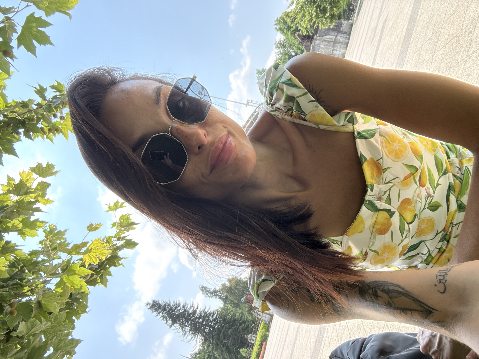 The media file is a selfie taken by a woman outdoors. She is wearing a yellow and white top with a lemon print and sunglasses. Her hair is brown and shoulder-length. She has tattoos on her arm. The background shows a blue sky with some clouds, green trees, and what appears to be a paved area. The lighting suggests it is daytime, likely sunny. The image is taken at a slightly upward angle. There is some visible Arabic text on her arm tattoo. The scene appears to be a park or an outdoor seating area, possibly in Pleven, Bulgaria, given the location context. The woman has a slight smile and seems relaxed.