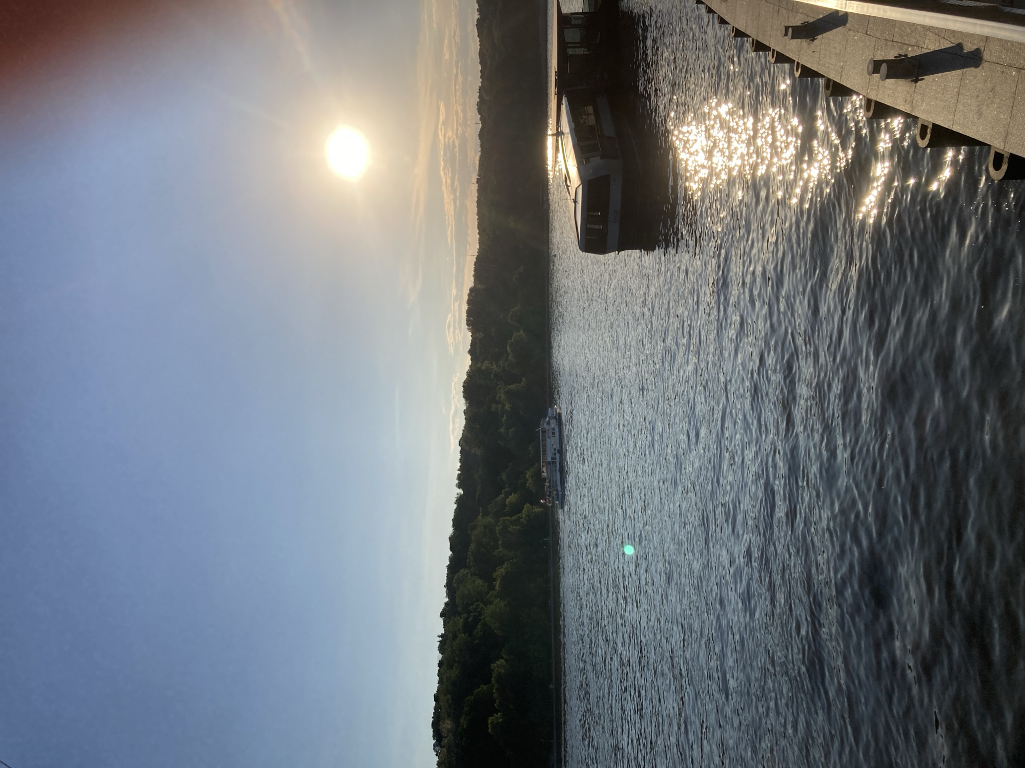 This is a photograph taken on the Moskva River in Moscow, Russia, likely during the golden hour of sunset. The sun is low in the sky, casting a warm glow and creating a shimmering reflection on the water.
In the foreground, to the right, is a section of a paved embankment with dark bollards. The water itself is a dark blue, with ripples reflecting the sunlight in a scattered pattern.
Further out on the river, there are two boats. One, a larger passenger boat, is docked or moored near the embankment on the right. It is a modern design with large windows and a flat roof. In the distance, on the left side of the river, a smaller passenger boat is navigating the water.
The background features a densely wooded hillside, indicative of a park or natural area along the riverbank. The sky is a clear, light blue with the sun positioned prominently in the upper right quadrant of the image. There are a few wispy clouds near the horizon. The overall atmosphere is serene and picturesque.