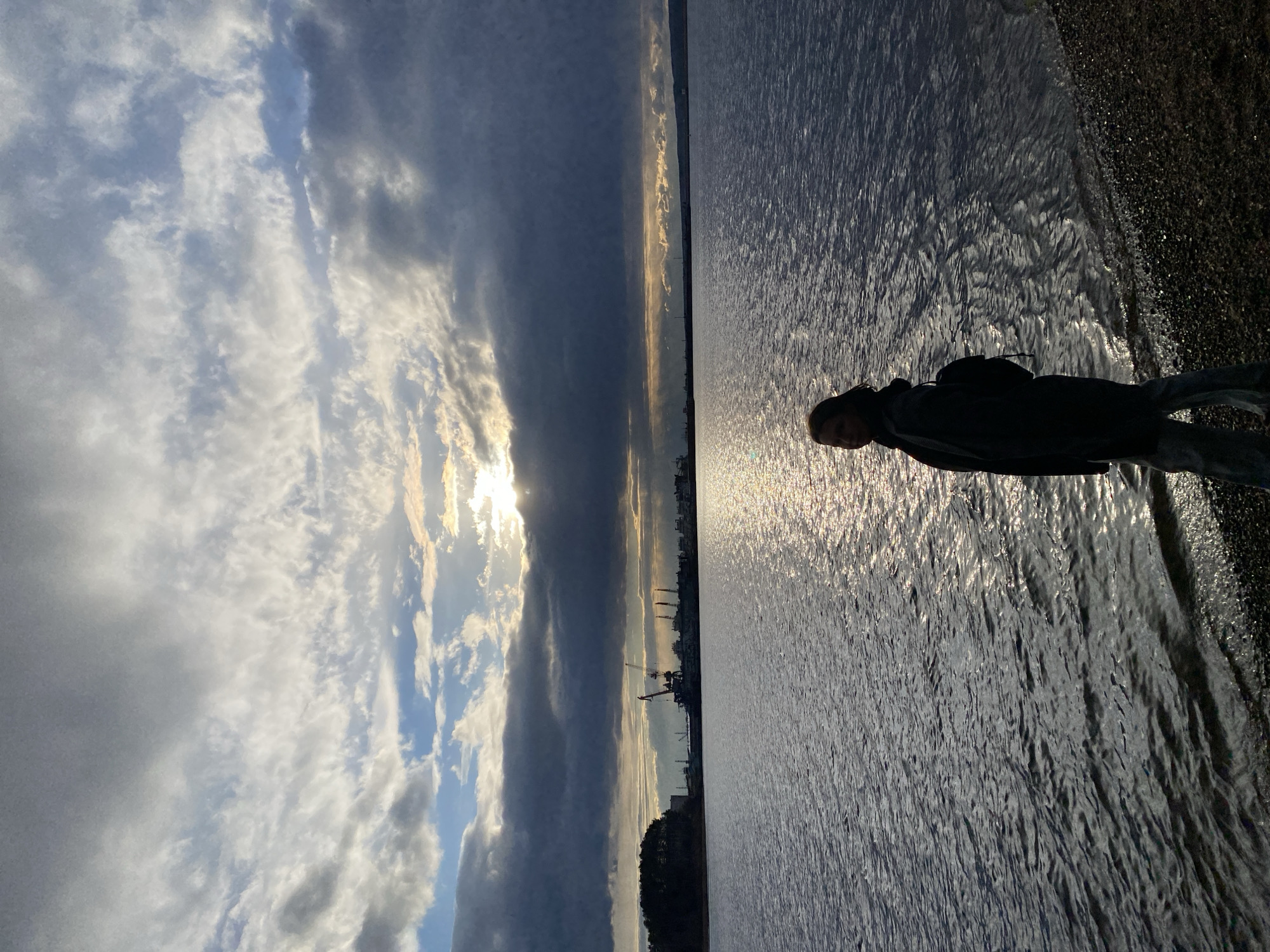 The image, oriented horizontally, presents a broad outdoor scene of a person standing by a large body of water under a dramatic sky.
In the upper half, the sky is filled with a mix of dense, dark clouds and brighter patches where sunlight breaks through. On the left side, the sun, partially obscured by clouds, shines intensely, creating bright highlights and golden hues within the cloud formations and on the water. Patches of blue sky are also visible amidst the clouds.
A distinct horizon line stretches across the middle of the image, separating the sky from the water. Along this distant horizon, the dark silhouettes of industrial structures, including cranes and buildings, are visible, suggesting a distant urban or port area.
The lower half of the image is dominated by a vast body of water, likely a river or reservoir, consistent with the location context of Chaikovsky, Russia. The water's surface is textured with ripples, and brilliant, shimmering reflections from the low-lying sun stretch across its left side, creating a striking contrast with the darker water elsewhere.
In the foreground, on the right side, a solitary figure stands facing slightly left, largely silhouetted against the bright reflections on the water. The person appears to be wearing a dark jacket or coat and trousers, possibly with a backpack. Their face, though mostly in shadow, shows some visible features due to the backlight. They stand on a dark, rocky or pebbly shore that occupies the extreme right edge of the image.
The scene captures the late afternoon or early evening, evidenced by the dramatic lighting and low sun position. The overall mood is contemplative, emphasized by the solitary figure and the serene yet powerful natural elements. No visible text is present in the image.