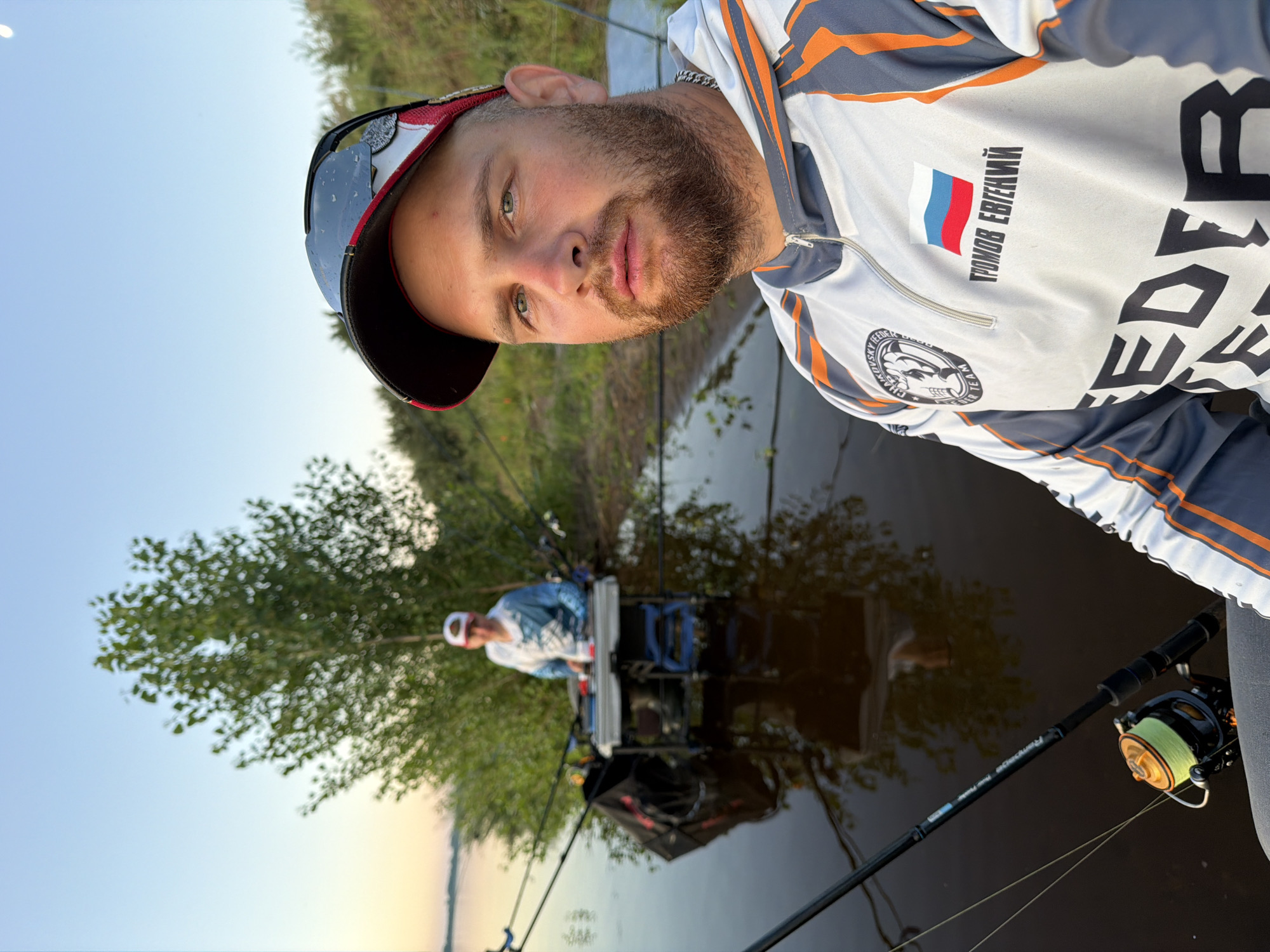 In Tarasovo, Russia, a man with a beard and a baseball cap is taking a selfie by the water. He is wearing a fishing shirt with the Russian flag and his name, "ГРОМОВ ЕВГЕНИЙ," printed on it. In the background, another person is fishing with multiple rods. The scene is set at dusk, with a tree and a body of water reflecting the sky. Fishing equipment, including rods and a tackle box, is visible.