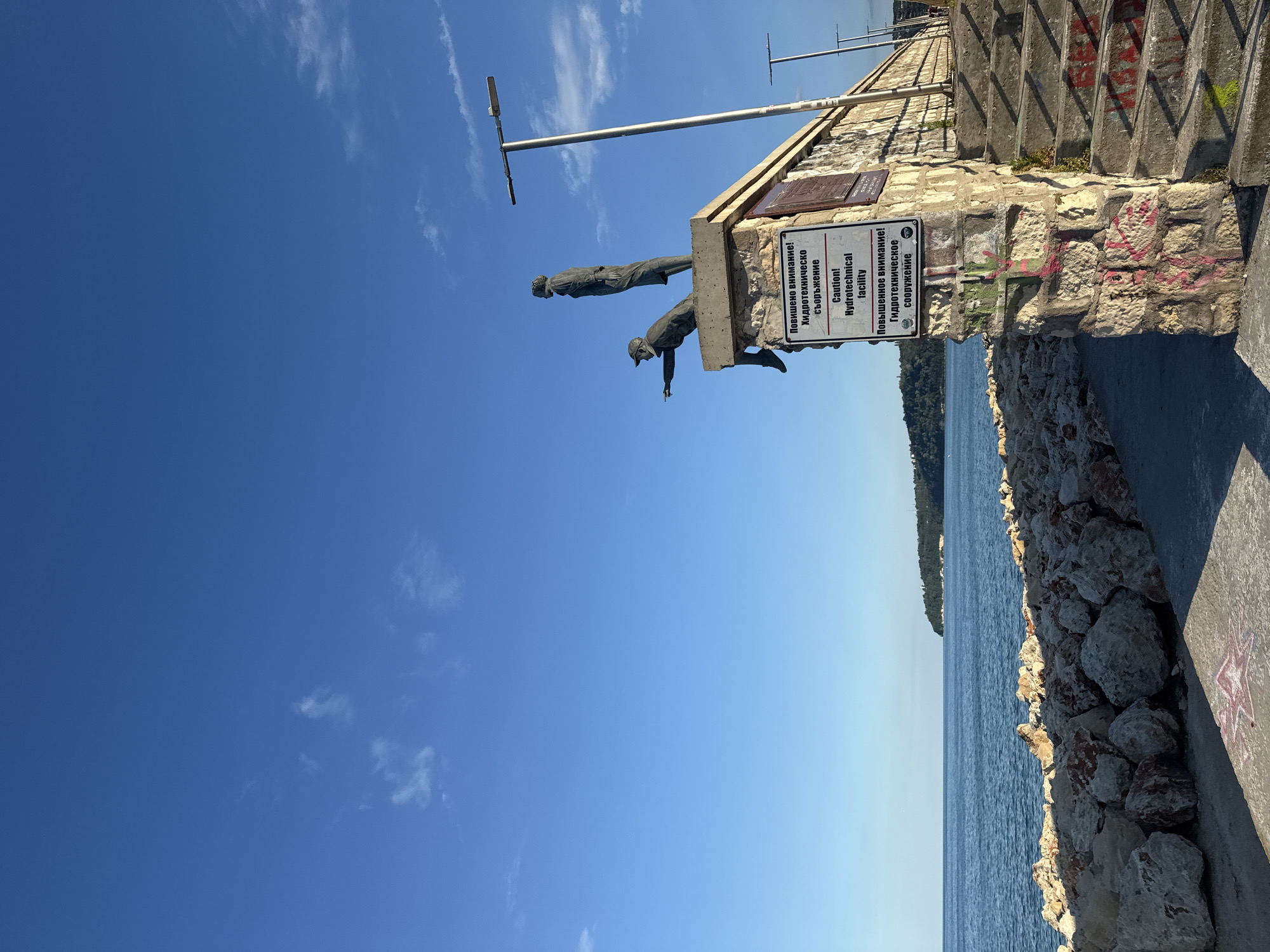 This image depicts a section of the Varna Sea Garden in Varna, Bulgaria, specifically the area near the Monument of the Ukrainian Cossacks. The scene is dominated by a blue sky with scattered white clouds. In the foreground, a stone structure serves as a base for two bronze statues of men. One statue, seated and pointing outward, is positioned on the left, while another standing statue is on the right. Below the statues, a sign is affixed to the stone wall, bearing bilingual text in Bulgarian and English. The English portion reads: "Caution! Hydrotechnical facility". Graffiti in various colors, predominantly pink and green, is visible on the stone base of the structure and on the concrete stairs leading upwards to the right.
To the left of the stone structure, a breakwater made of large rocks lines the edge of the sea. The sea itself is a deep blue, with a calm surface reflecting the sky. In the distance, a green, tree-covered coastline is visible under the bright sunlight, suggesting a clear day. Several lampposts with modern lighting fixtures are spaced along the walkway or embankment. The overall impression is of an outdoor public space, likely a popular tourist spot, with elements of both historical commemoration (the statues) and functional infrastructure (the hydrotechnical facility sign). The presence of graffiti indicates a degree of urban wear and tear on the otherwise well-maintained public area.