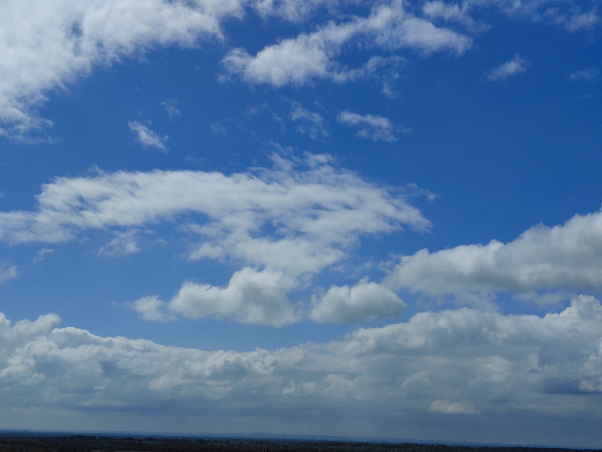 The image shows a wide expanse of blue sky with numerous white clouds of varying sizes and densities. The clouds are predominantly cumulus and stratocumulus formations, scattered across the sky at different altitudes. Some clouds are thin and wispy, allowing the blue sky to show through, while others are thicker and more opaque, casting subtle shadows. In the lower part of the image, a dark, hazy horizon line is visible, suggesting a landscape beneath the sky. This landscape appears to be a natural environment, possibly with vegetation or distant structures, but it is largely obscured by atmospheric haze and the framing of the image which focuses heavily on the sky. The overall impression is of a partly cloudy day. The provided information indicates the location as Novokuybyshevsk, Russia.