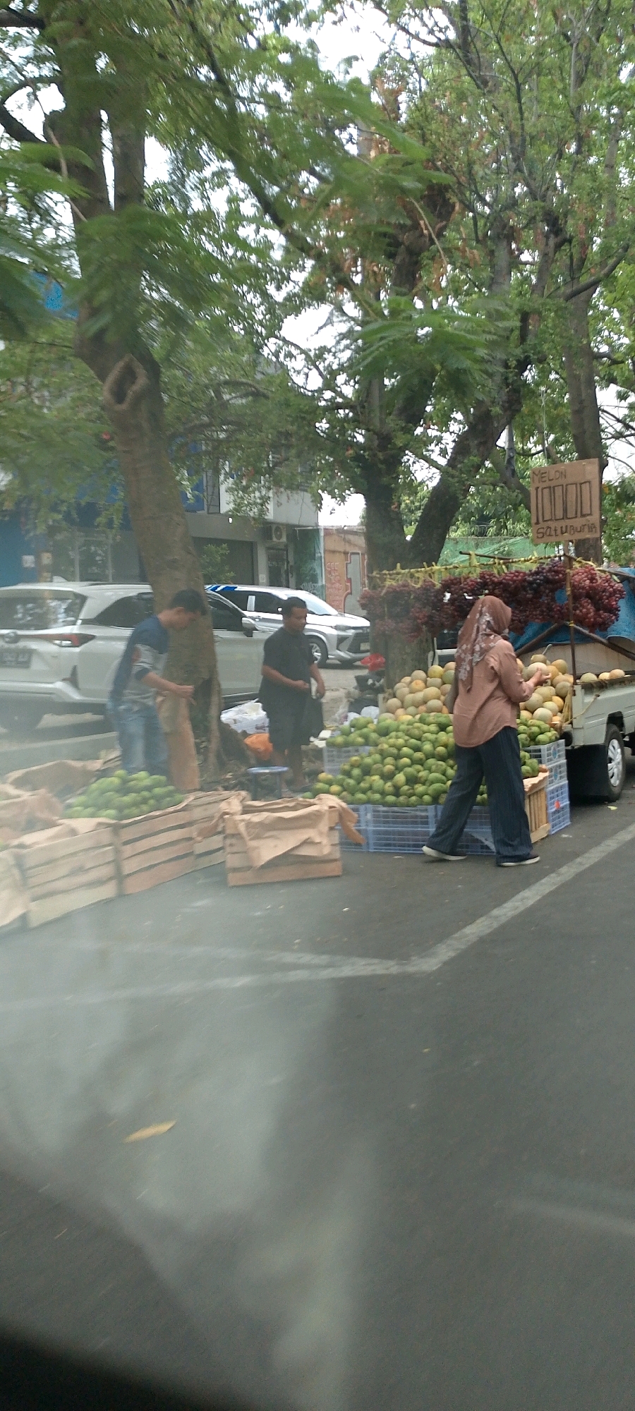 The image depicts a street scene with a fruit stand in what appears to be Bandung City, Indonesia. Several people are present, including a woman wearing a hijab and a woman in a tan colored hijab selecting a melon from a large pile. Two men are also visible, one in a blue shirt and jeans and the other in a black shirt. They are standing near several wooden crates filled with fruit. The fruit stand is situated on the side of a road with cars parked in the background, indicating an urban environment. Trees line the street, providing shade over the fruit stand. A sign reading "MELON 10000 SATUBURIR" is displayed above the fruit. The overall impression is of a bustling market scene in a tropical or subtropical climate.