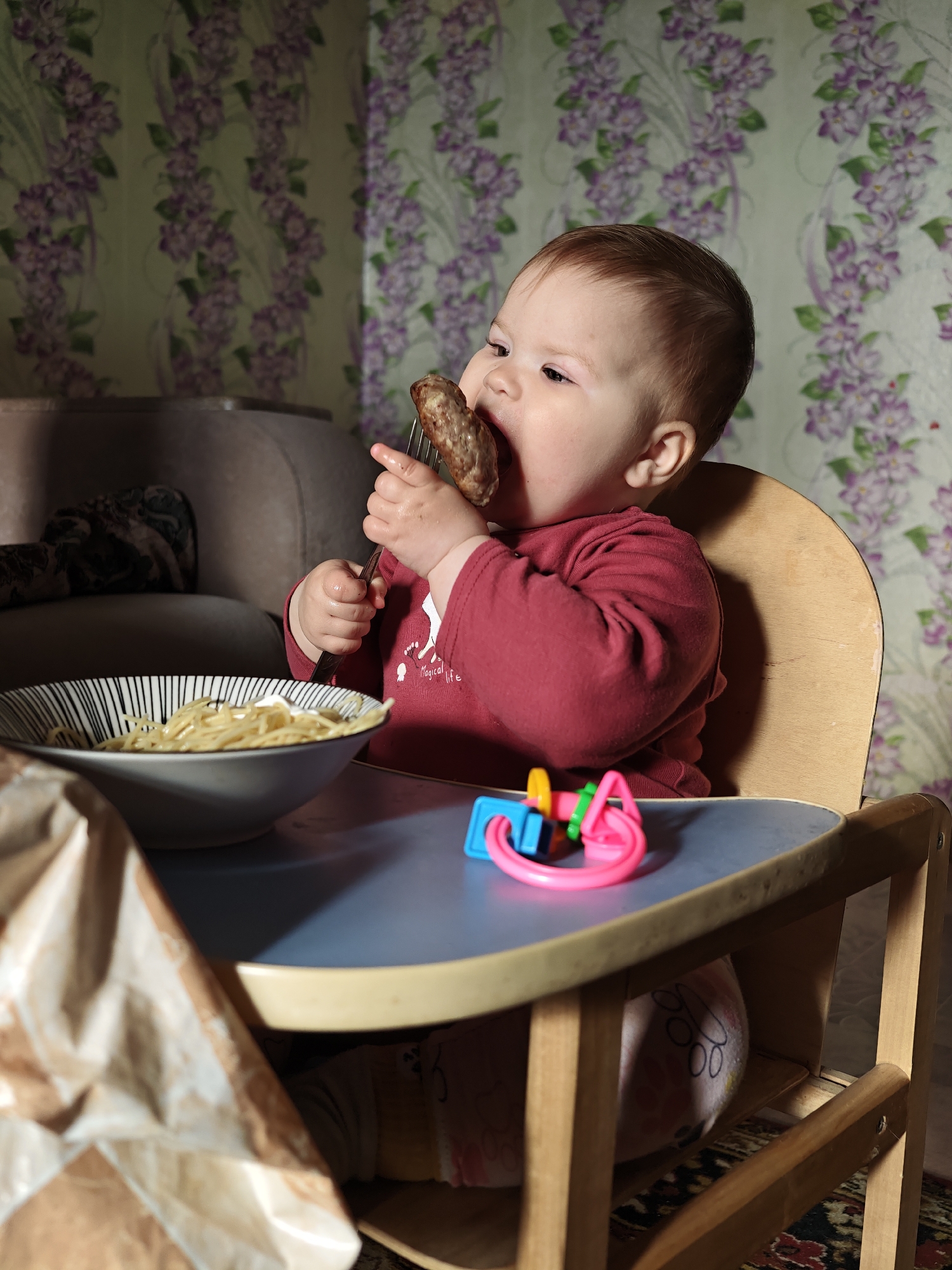 A baby in a high chair is eating a large piece of food, possibly a sausage, from a fork. There is a bowl of pasta on the table in front of the child, along with a colorful plastic toy. The baby is wearing a red long-sleeved shirt. The high chair is wooden with a blue table surface. The background features a floral wallpaper. A brown paper bag sits on the left side of the table. Part of a grey sofa is visible in the background. Location: Уральское, Russia.