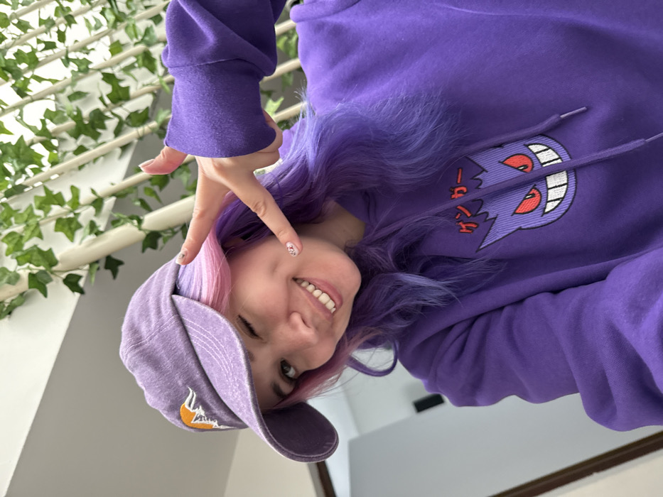 A young adult female is depicted in a close-up, chest-up shot within an indoor residential or commercial setting in the City of Zagreb, Croatia. She has light skin, winks with her right eye, and displays a broad, toothy smile. Her hair is dyed in a split style, appearing pink on the viewer's left side and purple on the viewer's right, styled with waves. She wears a faded purple baseball cap with an embroidered orange and white mountain and sun/moon motif, oriented slightly to her left. She is dressed in a purple hooded sweatshirt featuring an embroidered graphic of the Pokémon character Gengar, complete with red eyes and prominent fangs, positioned below Japanese text reading "ゲンガー" (Gengar). Her left hand is raised to her face, forming a peace sign with her index and middle fingers extended. Her fingernails exhibit a subtle patterned design. The background includes white vertical railings adorned with green artificial ivy, suggesting a staircase or balcony structure, alongside a plain light-colored wall.